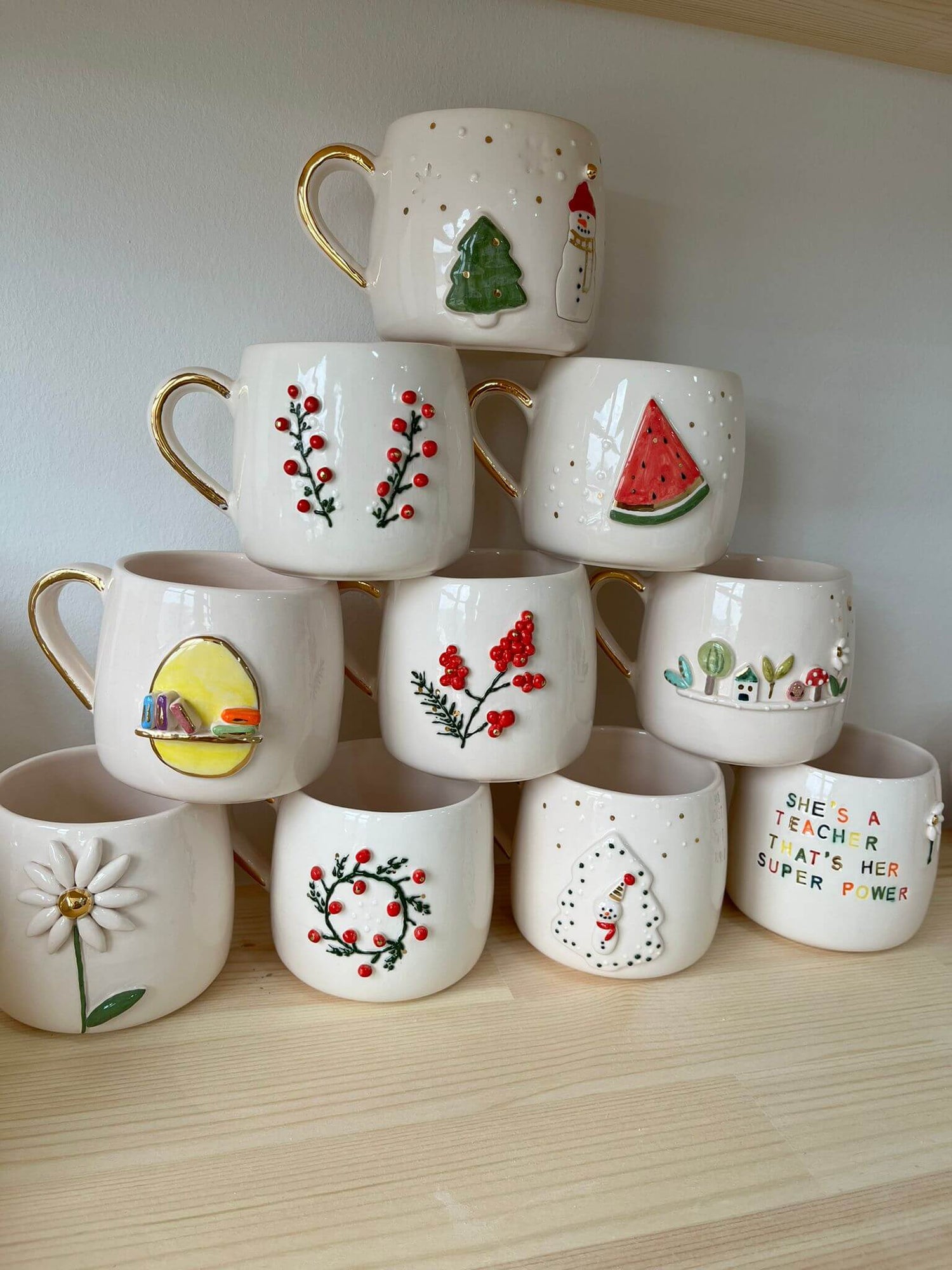 Handmade ceramic mugs featuring festive and playful designs, including holiday themes, watermelon slices, daisies, and a teacher's appreciation message, stacked neatly on a wooden shelf.