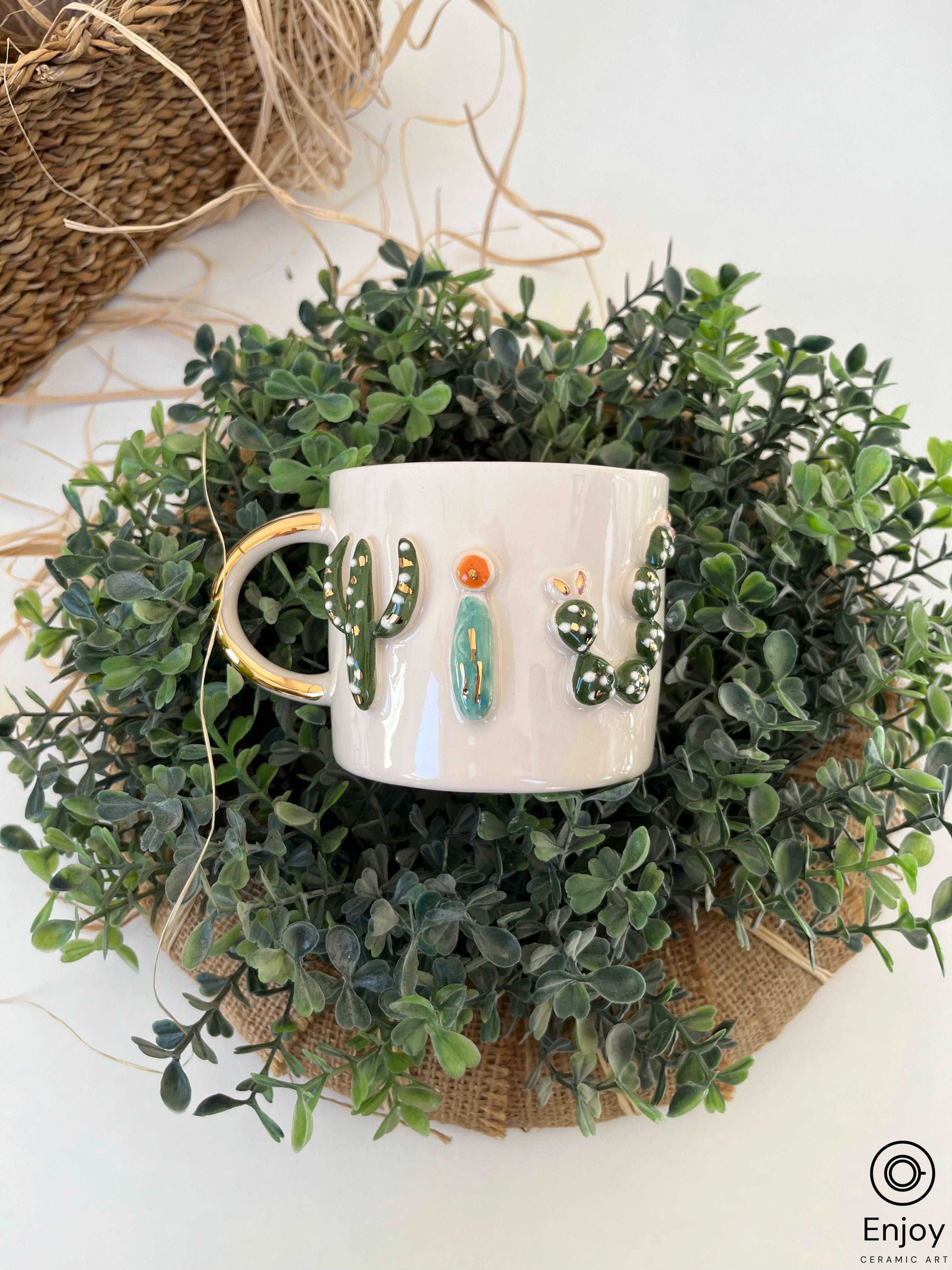 a white ceramic mug with a glossy gold handle, nestled in lush greenery. The mug is embellished with a raised, hand-painted cactus design in vibrant colors, offering a touch of nature's beauty to the setting.