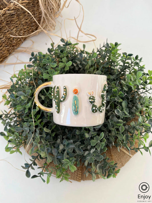 a white ceramic mug with a glossy gold handle, nestled in lush greenery. The mug is embellished with a raised, hand-painted cactus design in vibrant colors, offering a touch of nature's beauty to the setting.