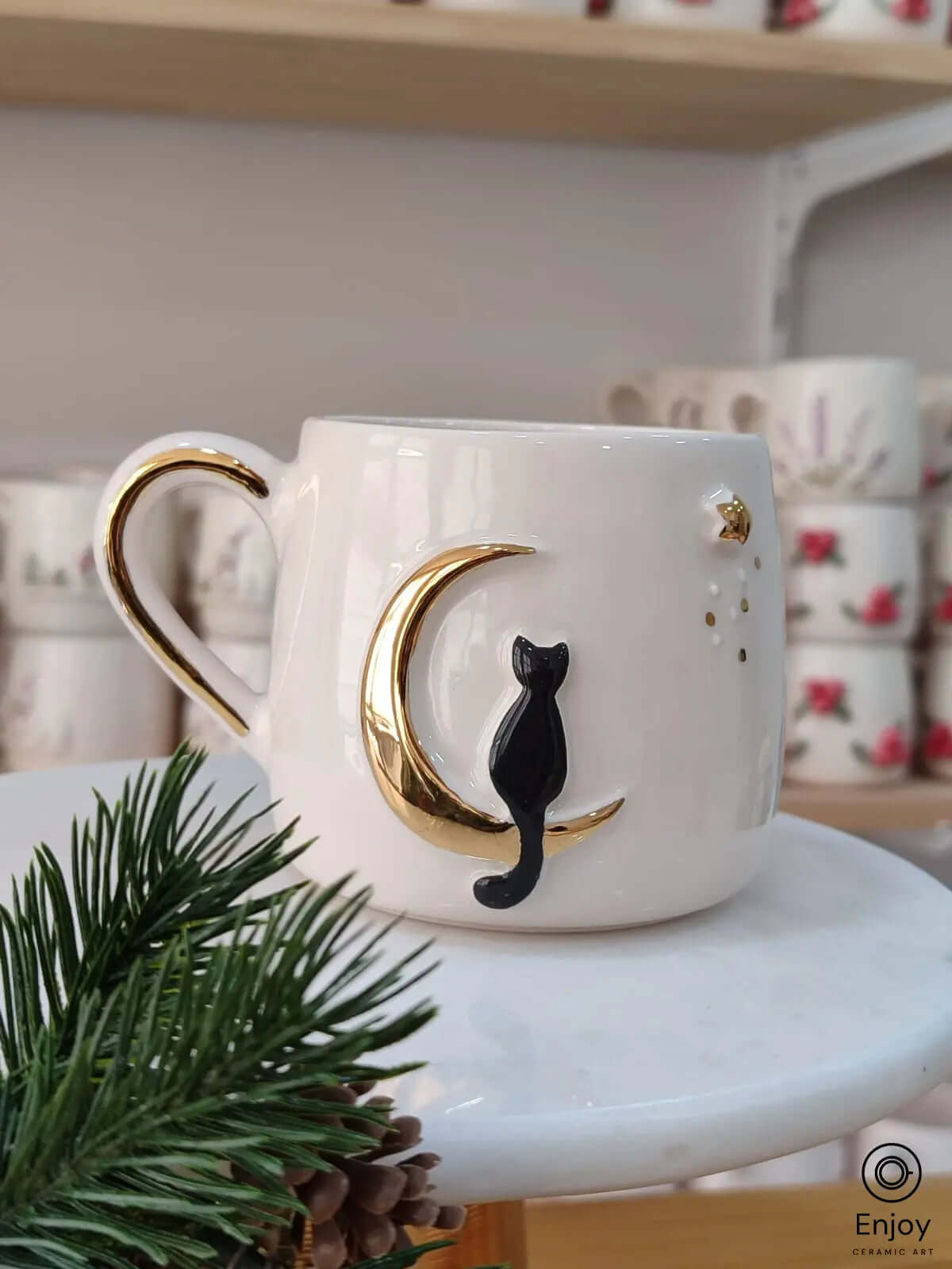 Handmade ceramic mug featuring a black cat sitting on a golden crescent moon, adding a mystical touch to your beverage experience, displayed on a marble stand with holiday accents.