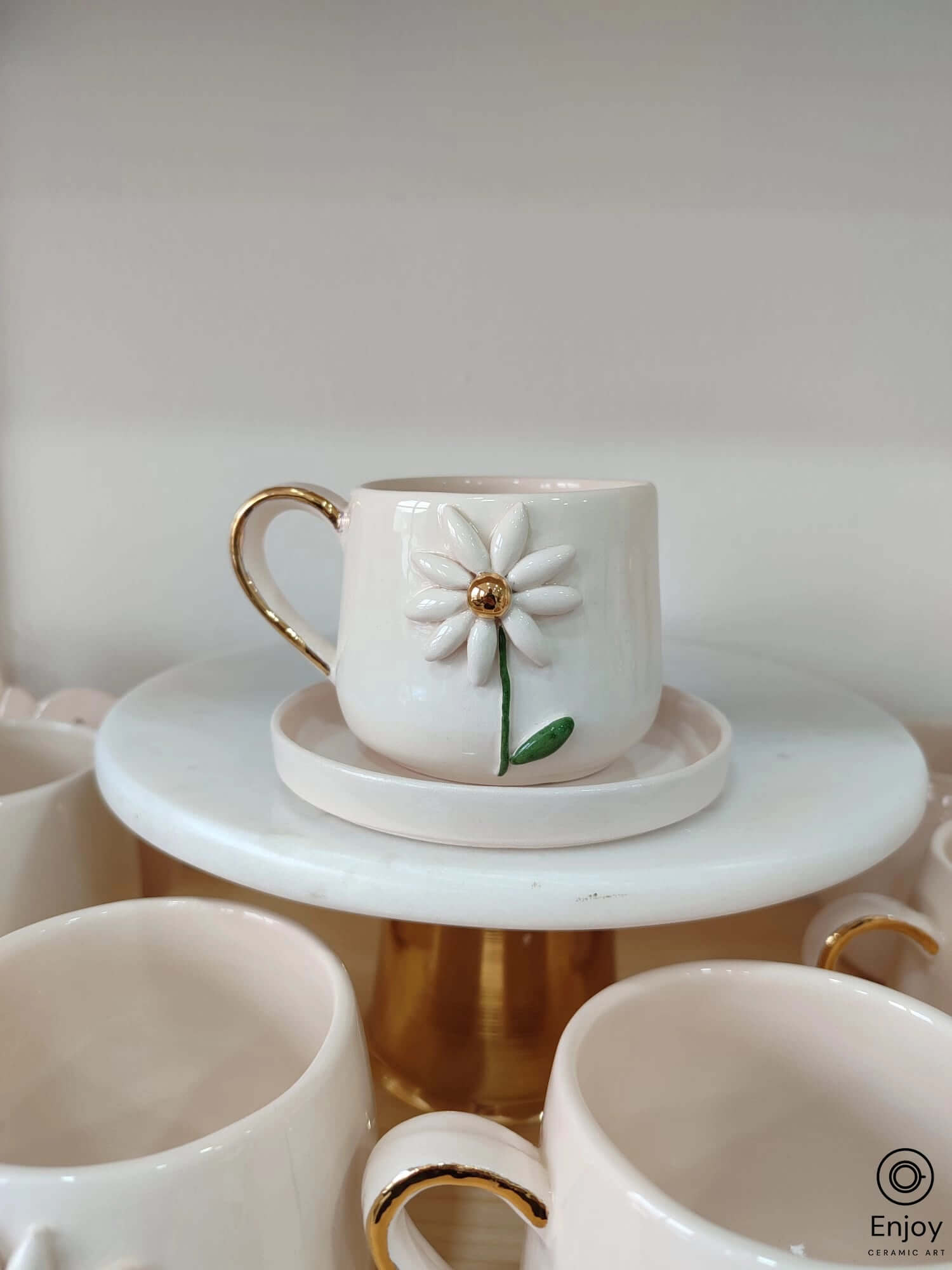 A white ceramic mug featuring a 3D daisy with a gold center and handle, resting on a matching saucer atop a golden pedestal.