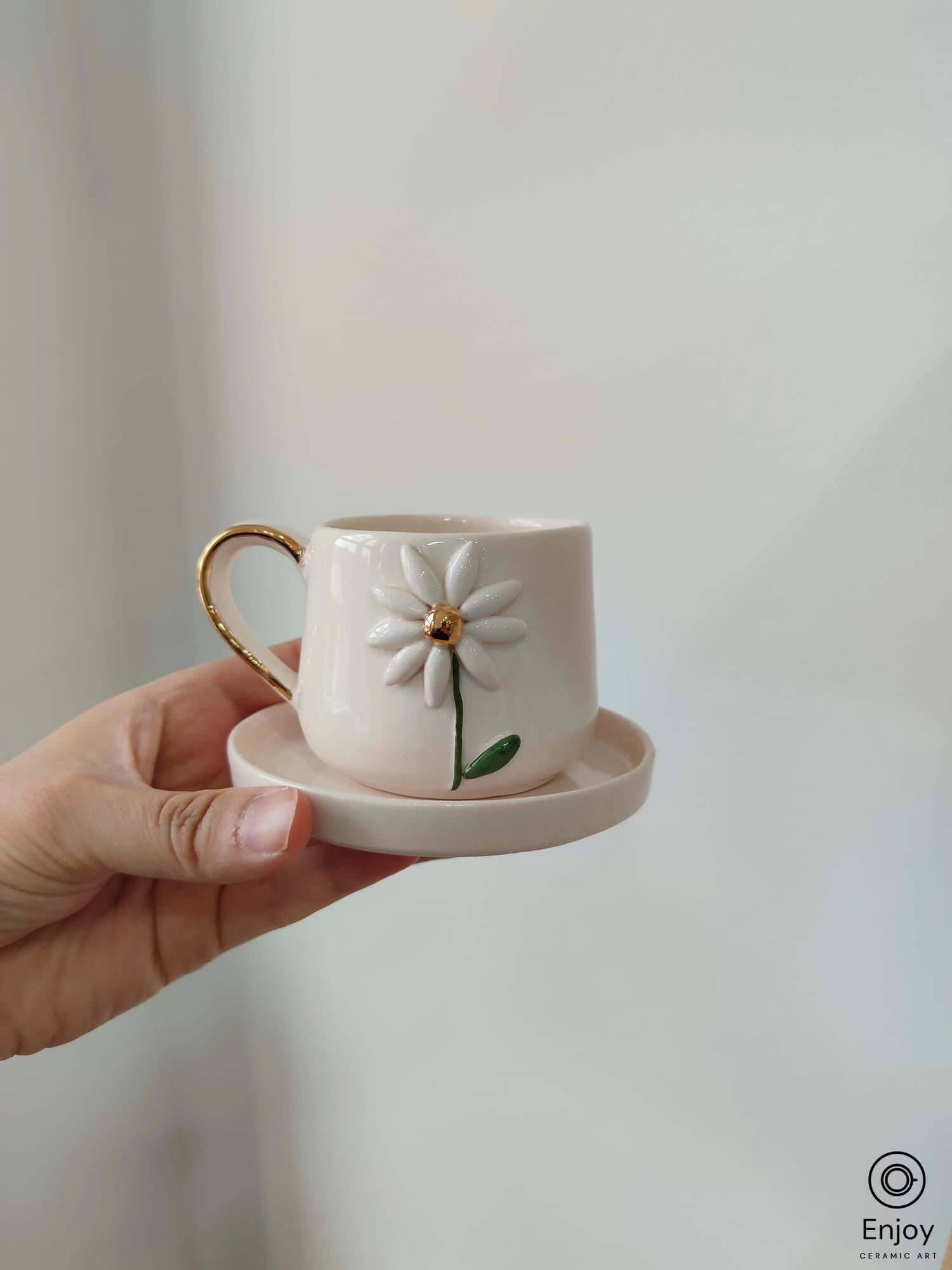 A delicate white ceramic mug with a 3D daisy design, gold handle, and matching saucer, held in hand.