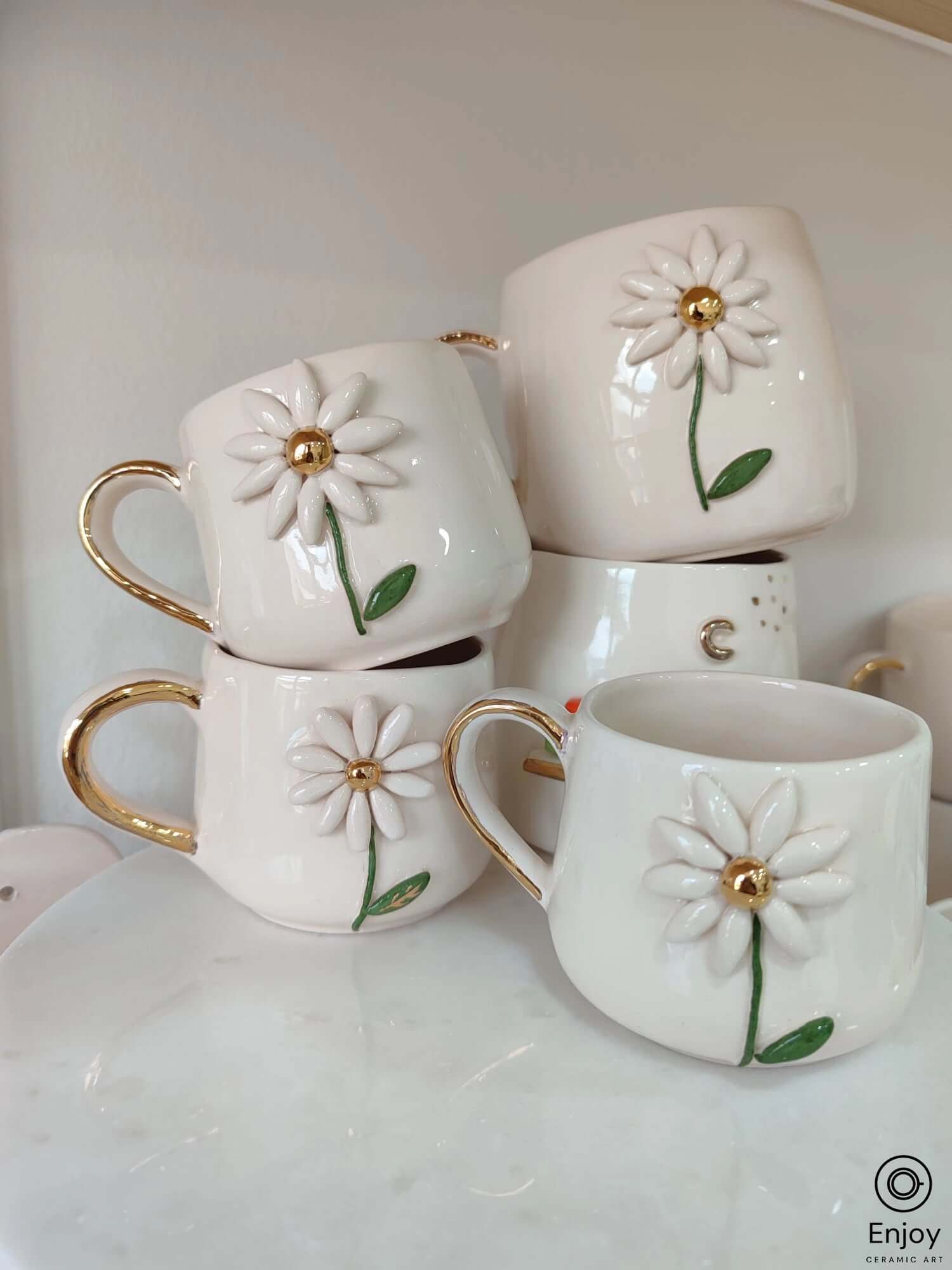 A collection of white ceramic mugs adorned with 3D daisy designs and gold handles, stacked neatly.