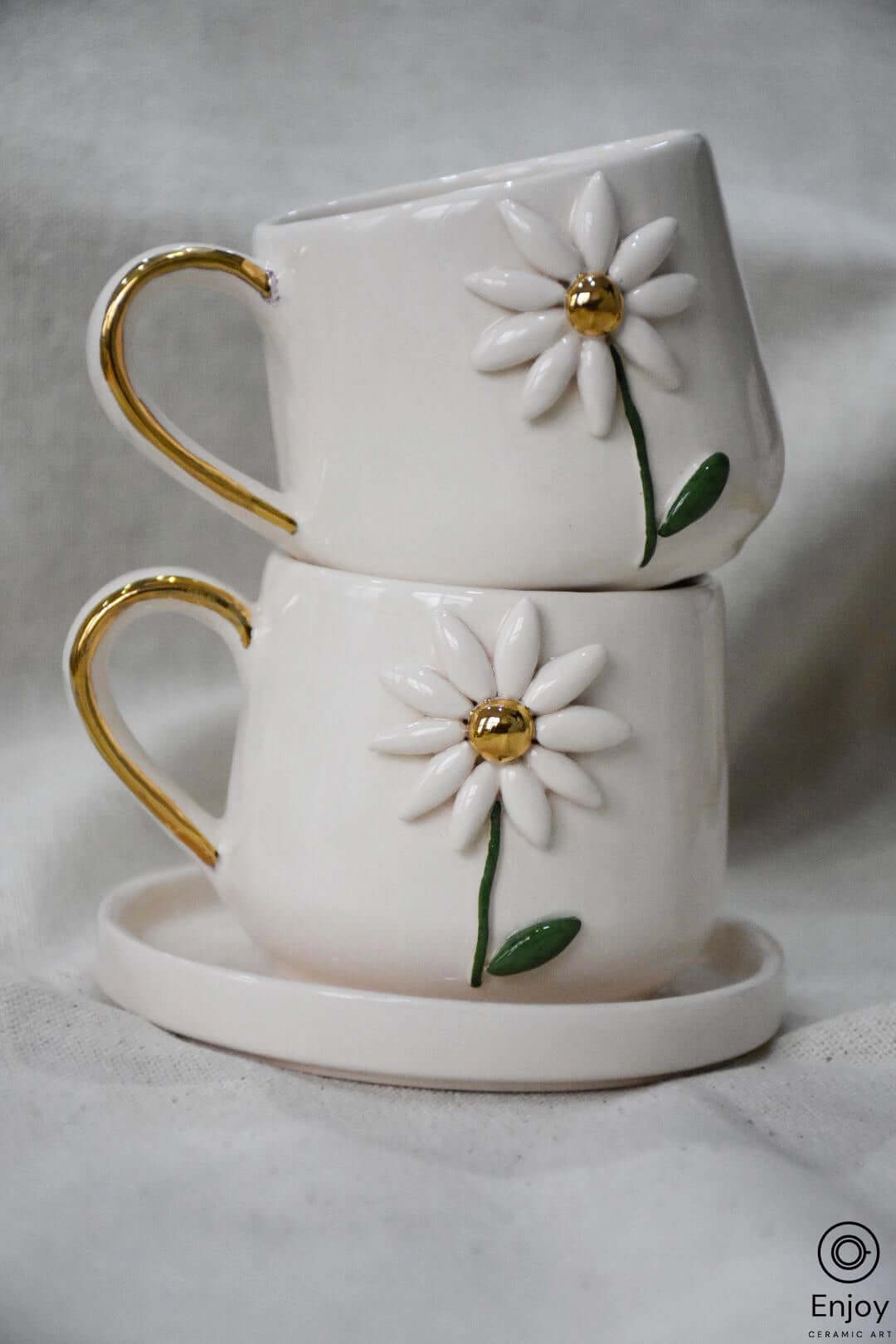 Two white mugs adorned with 3D daisy designs and gold handles, stacked on a matching saucer.