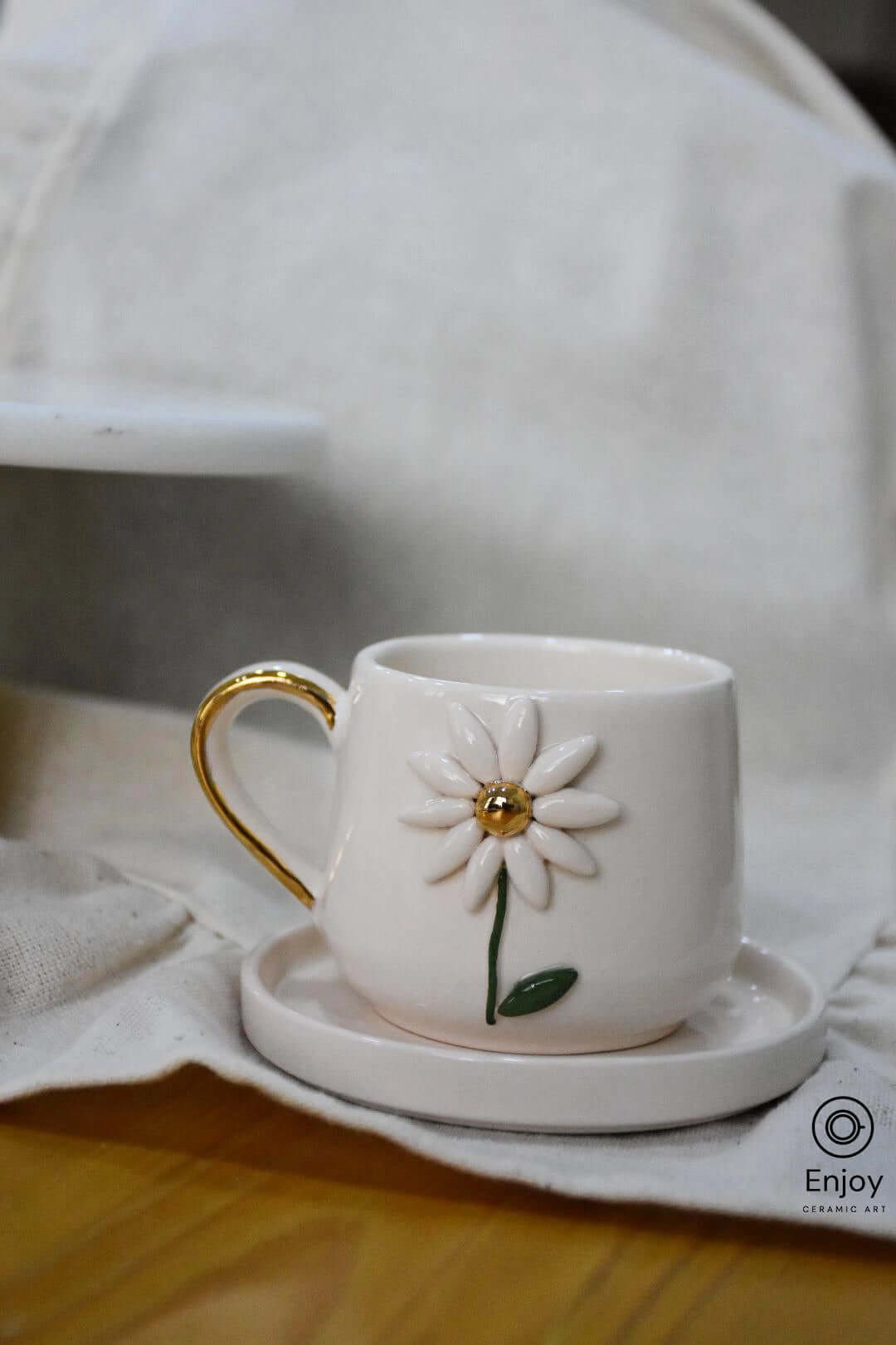 A white ceramic cup with a 3D daisy design and gold handle sits on a matching saucer against a soft linen background.