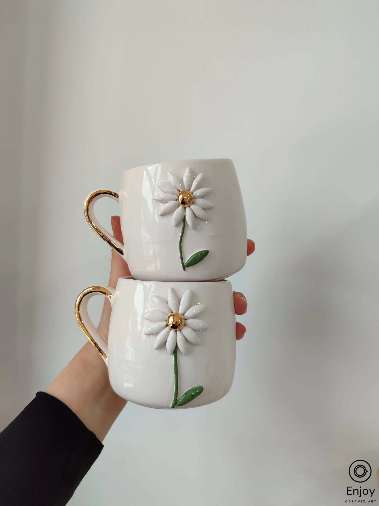Two white ceramic mugs featuring a 3D daisy design with gold handles, stacked together against a plain background.
