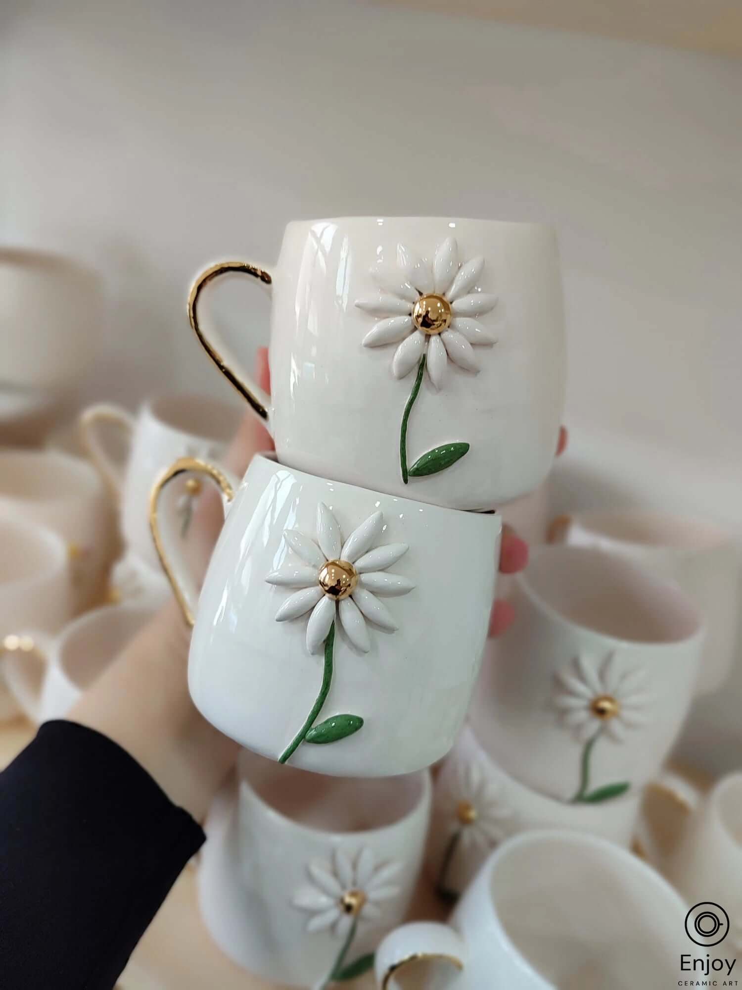 Two white ceramic mugs with daisy designs and gold handles, stacked and displayed among other similar mugs in the background.