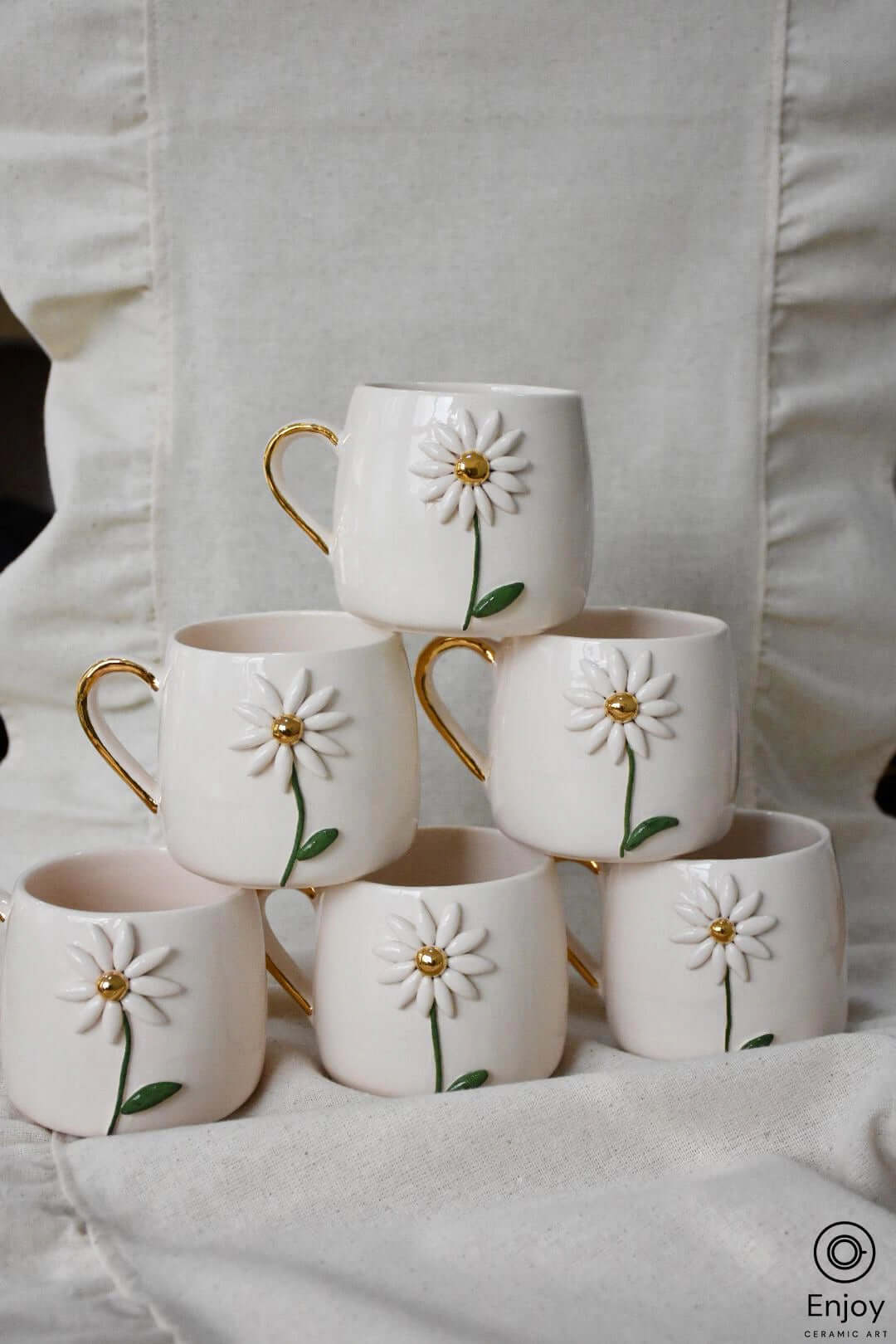 Six white ceramic mugs with daisy designs and gold handles stacked in a pyramid formation against a linen backdrop.