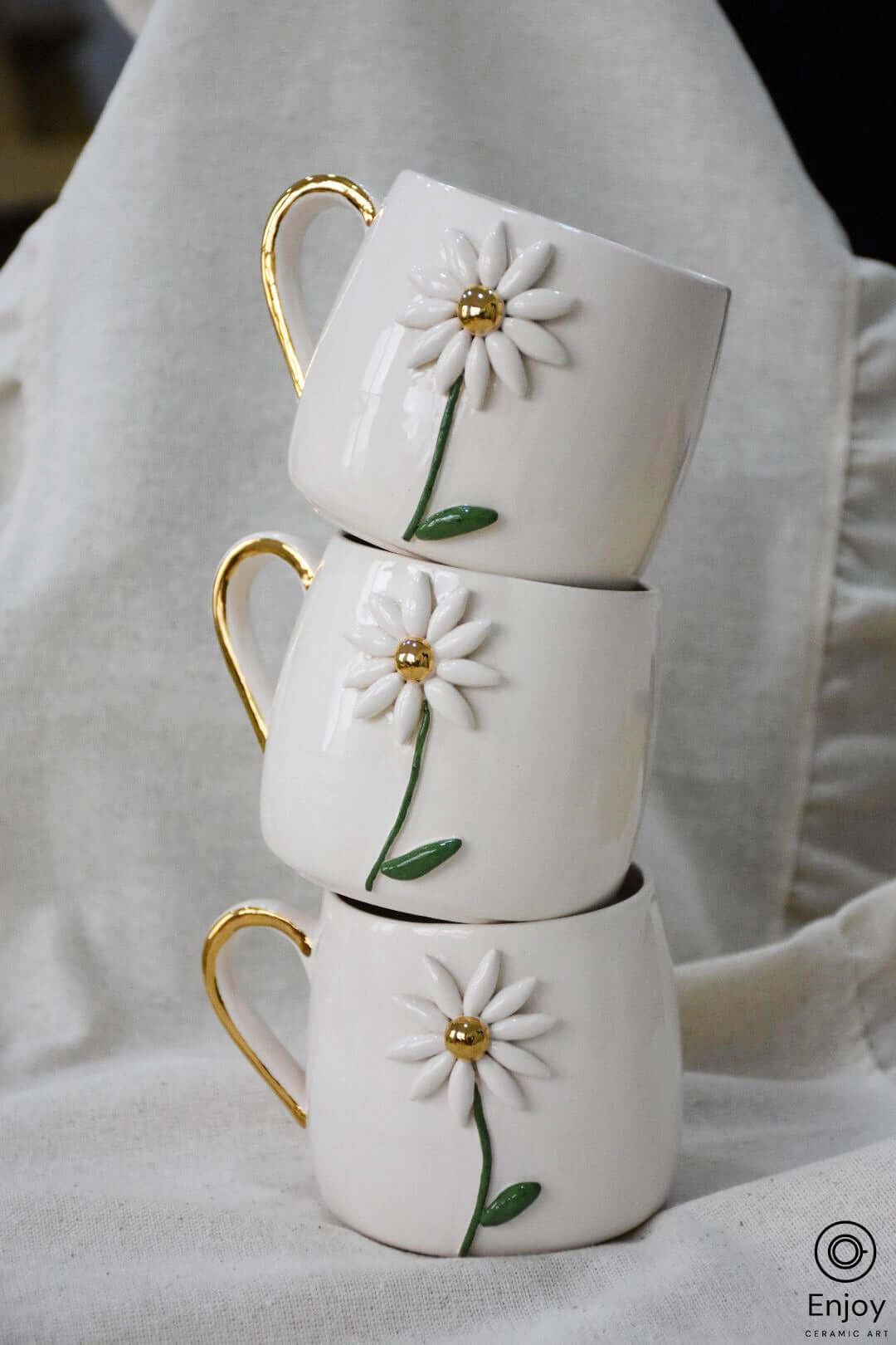 Three white ceramic mugs adorned with daisy designs and gold handles, stacked neatly on a linen cloth background.