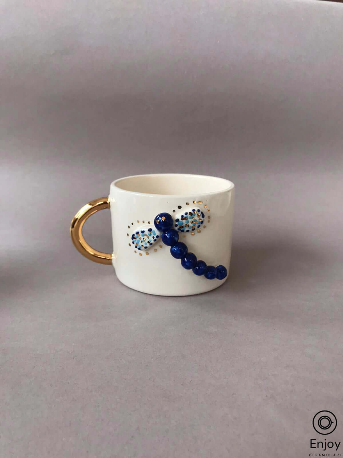 Handcrafted 'Blue Dragonfly Handmade Ceramic Coffee Mug' with a gold handle, displayed on a lavender background for a simple and elegant presentation.
