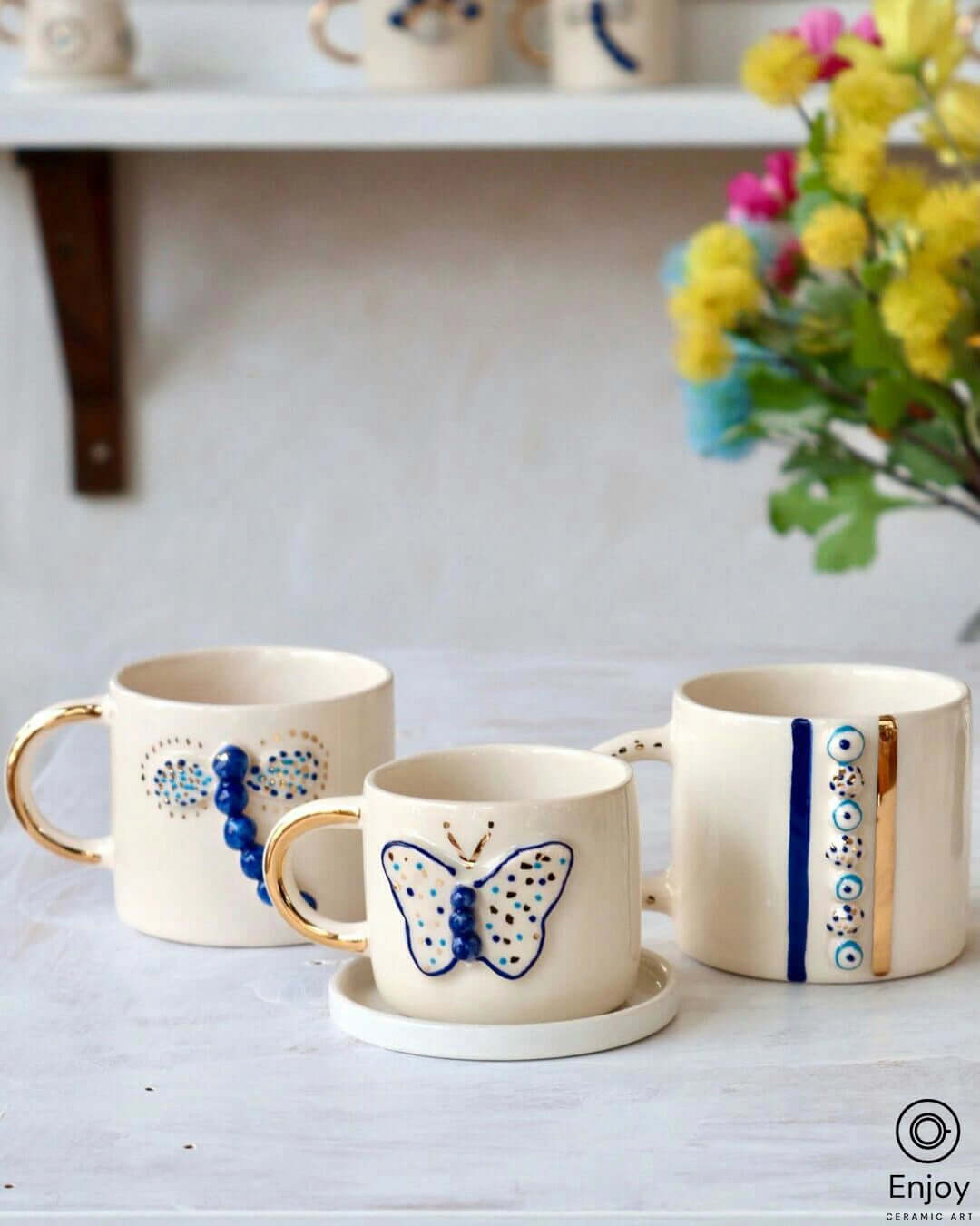 Three handmade ceramic coffee mugs with gold handles and blue bead dragonfly designs on a rustic shelf, complemented by vibrant spring flowers in the background for a fresh, artistic display.