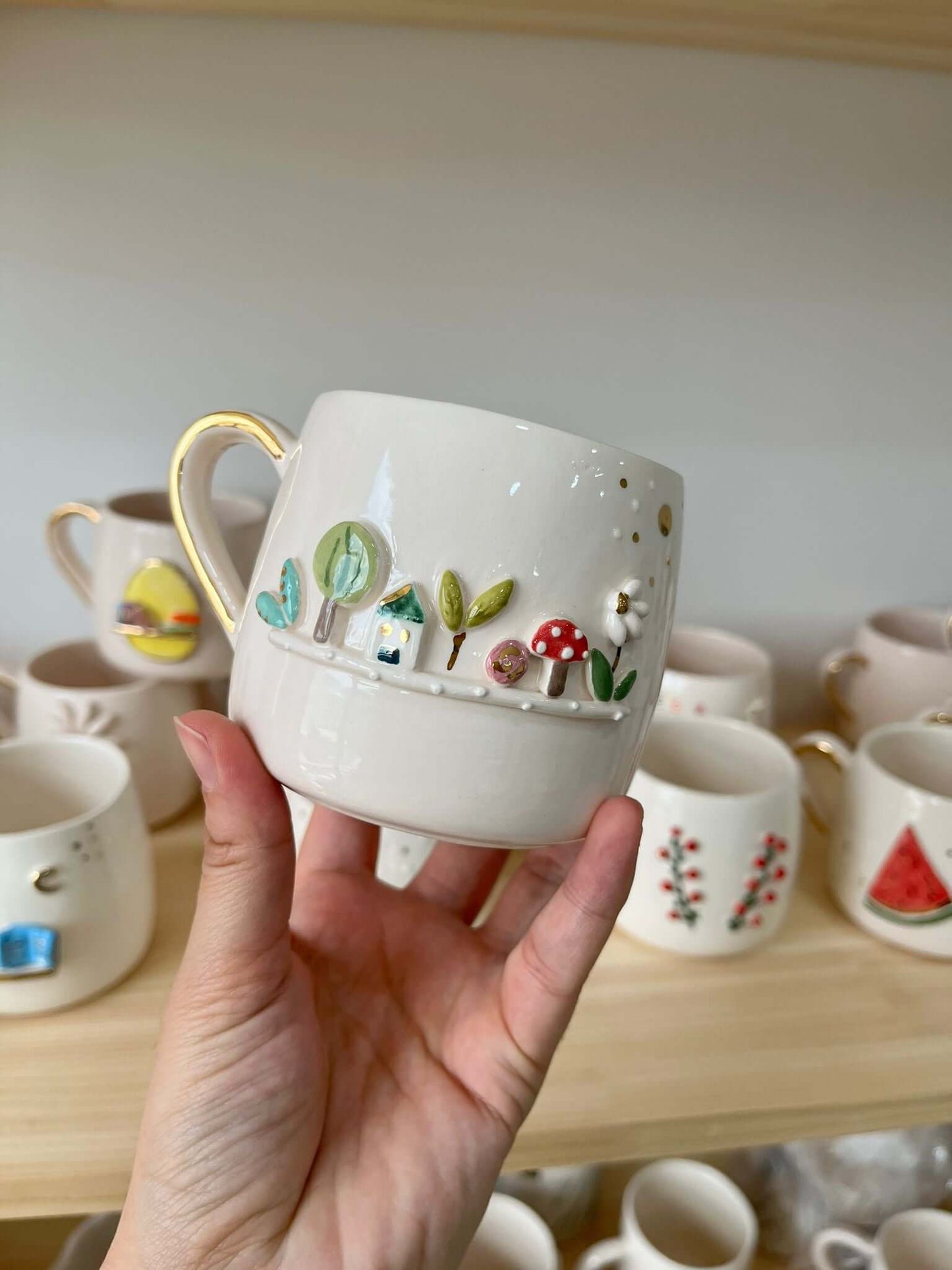 A handcrafted white ceramic mug with a 3D garden scene featuring trees, mushrooms, and a house, held in front of a display shelf.
