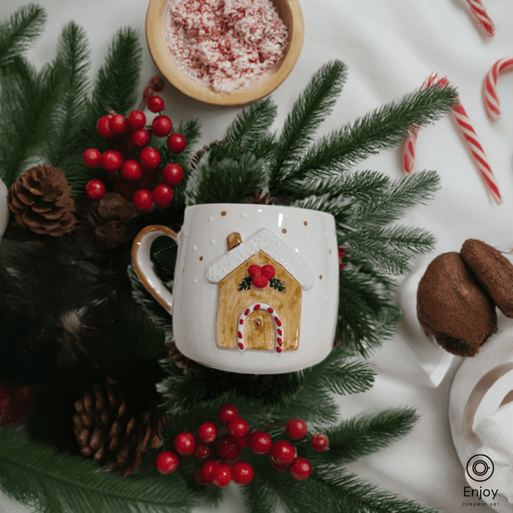 A festive ceramic mug featuring a cozy gingerbread house design, surrounded by pine branches, red berries, and candy canes, creating a warm holiday scene.