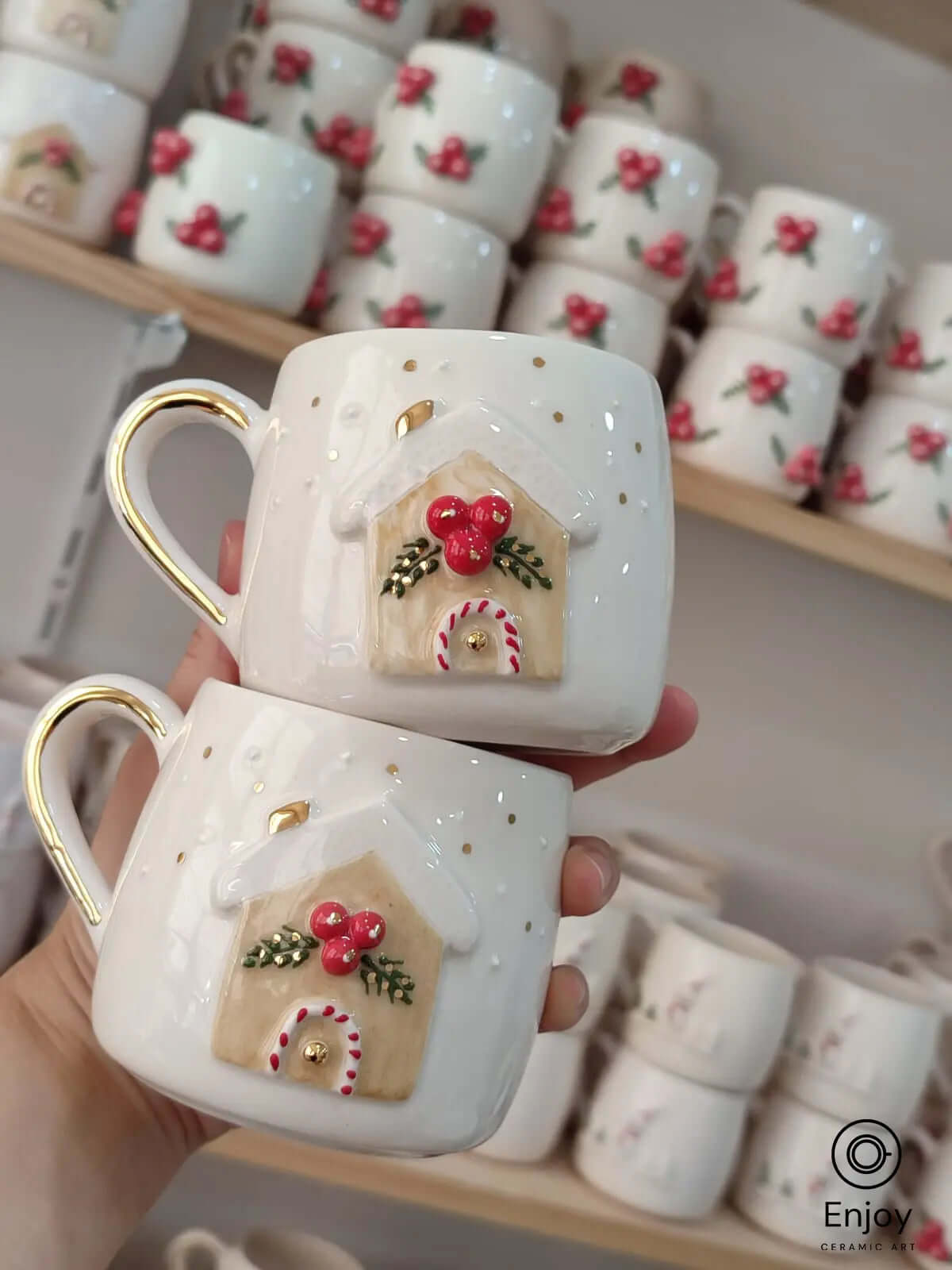 Handmade ceramic mug adorned with a festive gingerbread house design, complete with bright red berries, evergreen sprigs, and snowy accents. The golden handle adds an elegant touch, making it perfect for cozy holiday moments. Ideal for sipping hot cocoa by the fireplace or gifting to spread Christmas cheer.