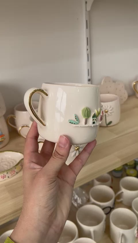 A handcrafted white ceramic mug with a 3D garden scene featuring trees, mushrooms, and a house, held in front of a display shelf.