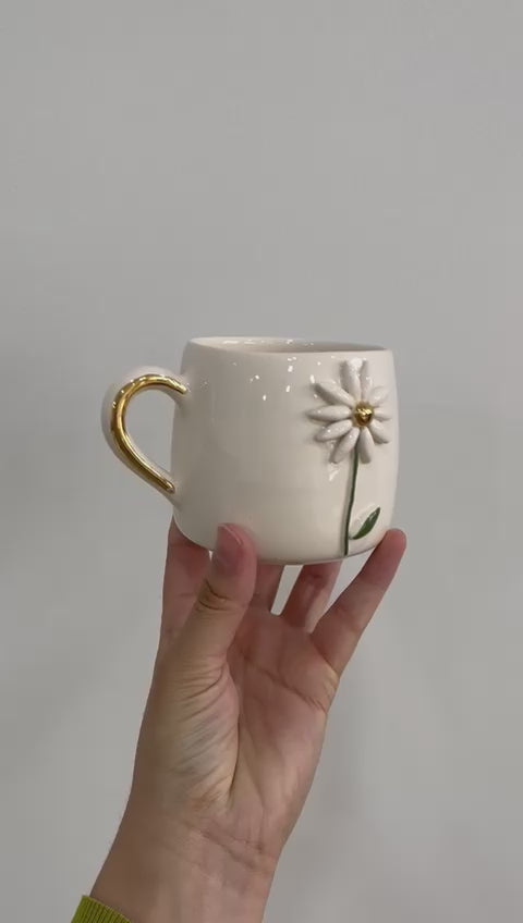 white ceramic mug featuring a 3D daisy design with gold handles,  against a plain background.