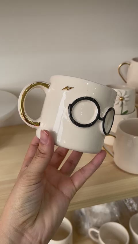 A hand gently rotates a ceramic mug featuring bold round glasses and a golden handle, set against a wooden shelf background, showcasing the unique design from different angles.