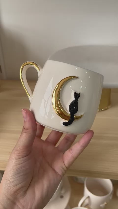 Hand gently rotating a ceramic mug with a black cat perched on a golden crescent moon, set against a wooden shelf backdrop.