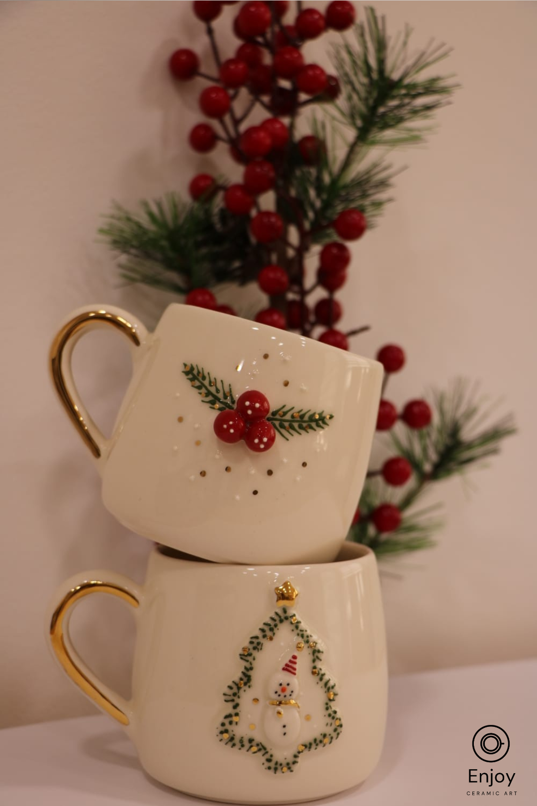Two stacked mugs with Christmas designs against a blurred background of red berries and greenery.