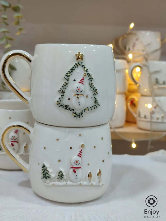 Two handmade ceramic mugs featuring festive 3D designs: a snowman framed in a Christmas tree and a snowy scene with a snowman, both with gold handles.