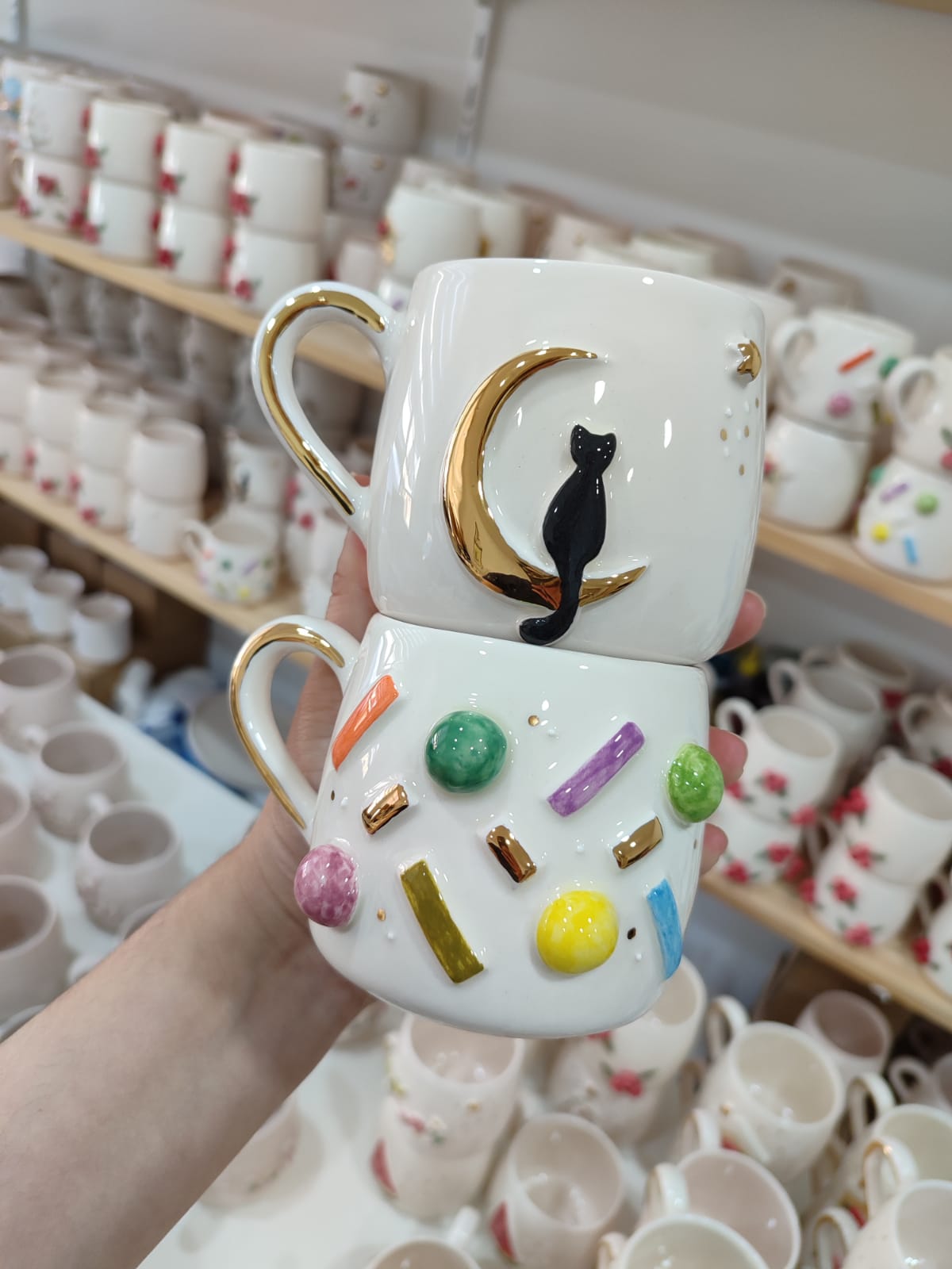 A pair of handmade ceramic mugs featuring a black cat on a golden crescent moon and vibrant 'sprinkles' design, stacked together in front of a display filled with more mugs.