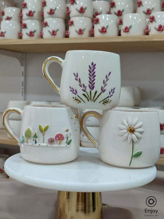 Set of three handmade ceramic mugs featuring daisy, lavender, and fairy garden designs, each with gold handles and detailing.