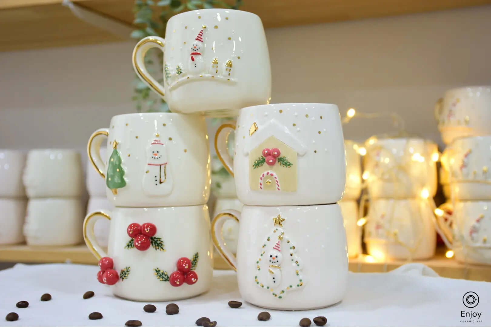 Set of five handmade ceramic Christmas mugs featuring festive designs: snowman, Christmas tree, gingerbread house, winter berries, and snowman in a tree frame, each with a gold handle.