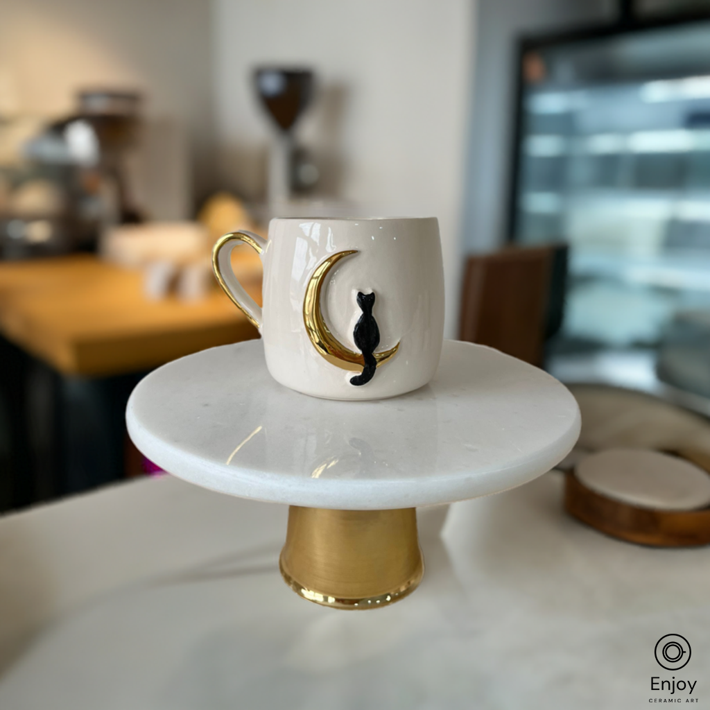 Handmade ceramic mug featuring a black cat perched on a golden crescent moon, displayed on a marble stand in a cozy cafe setting.