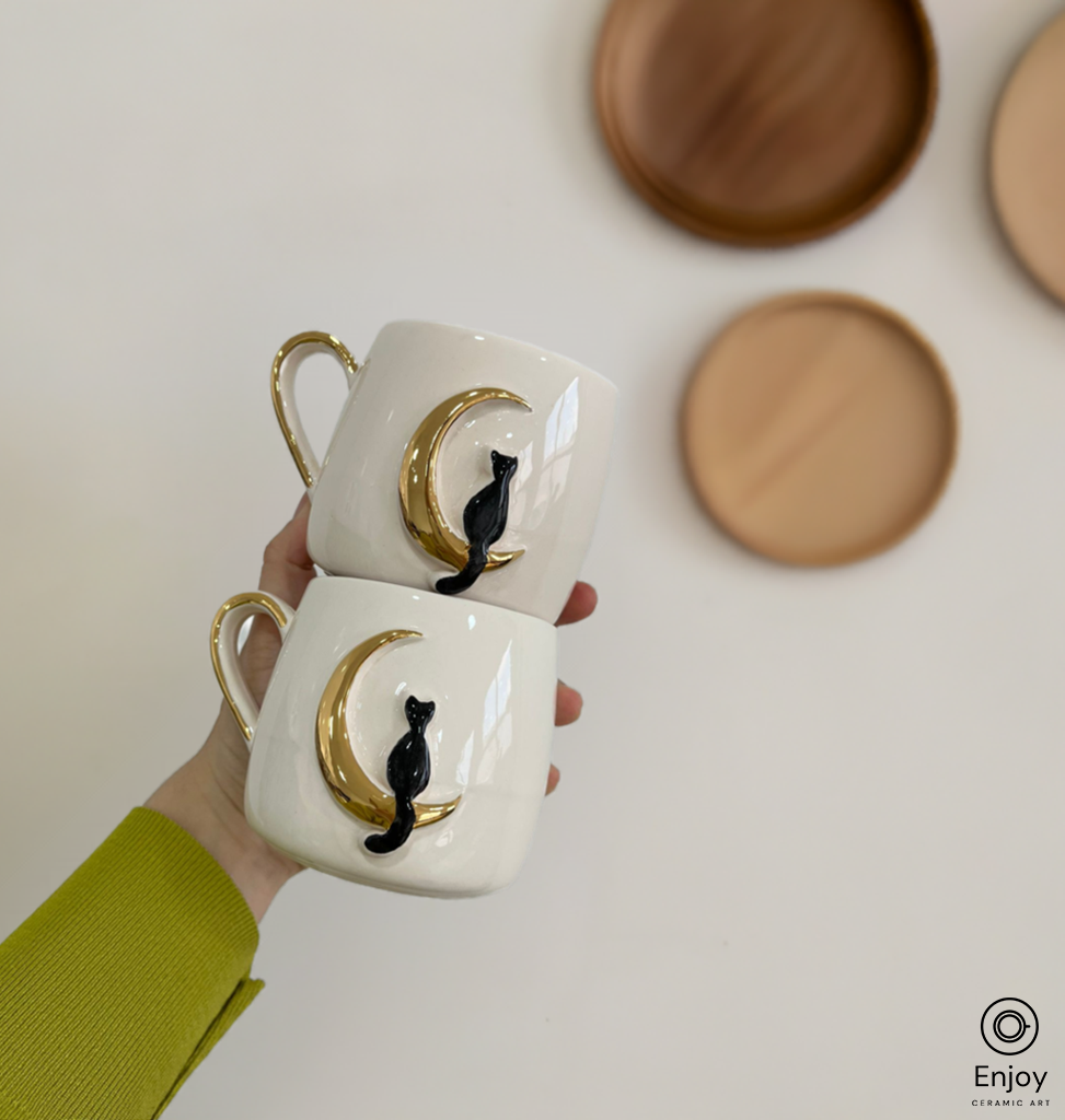 Hand holding two stacked ceramic mugs, each featuring a black cat perched on a golden crescent moon against a clean white background.