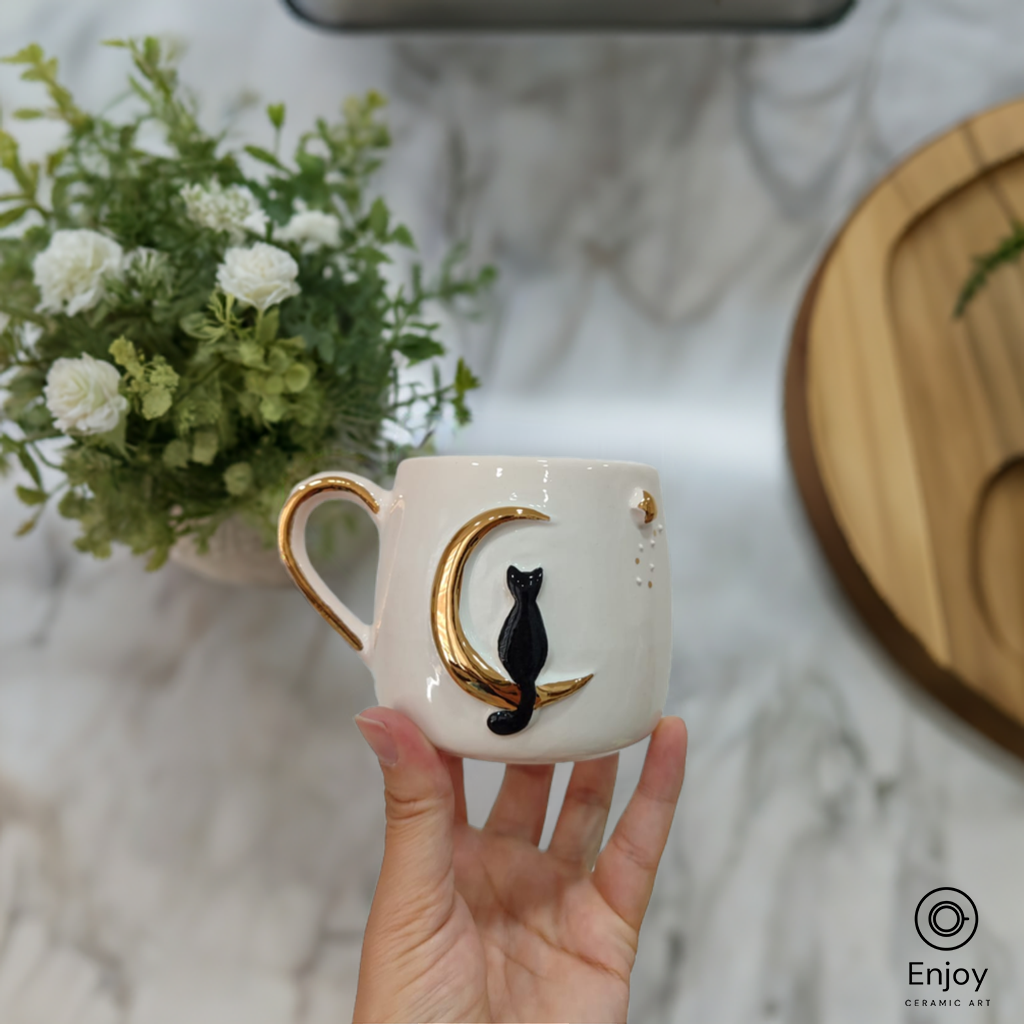 Hand holding a handmade ceramic mug featuring a black cat sitting on a golden crescent moon, with a floral background.