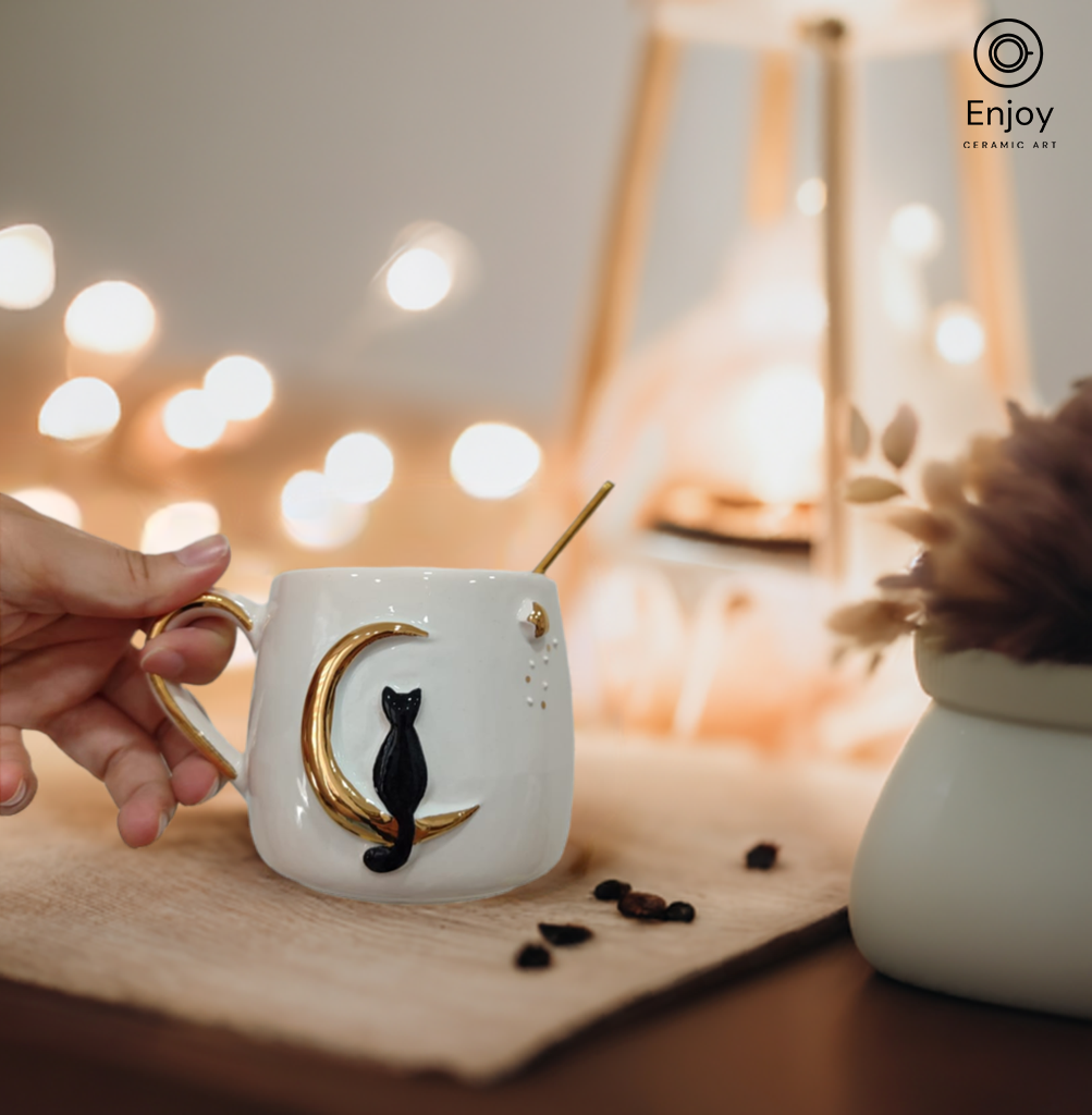 Hand reaching for a ceramic mug featuring a black cat on a golden crescent moon, with warm ambient lights in the background.