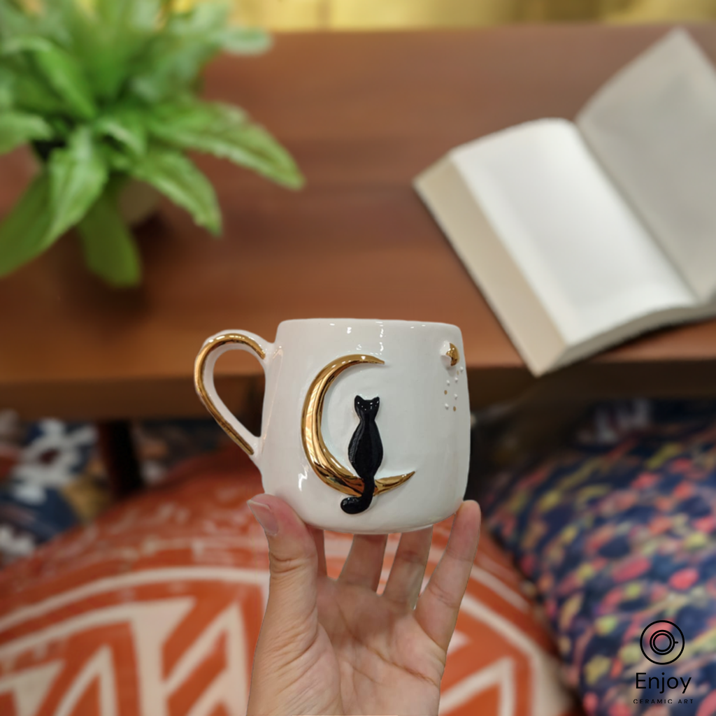 Hand holding a ceramic mug with a black cat on a golden crescent moon, cozy setting with an open book.