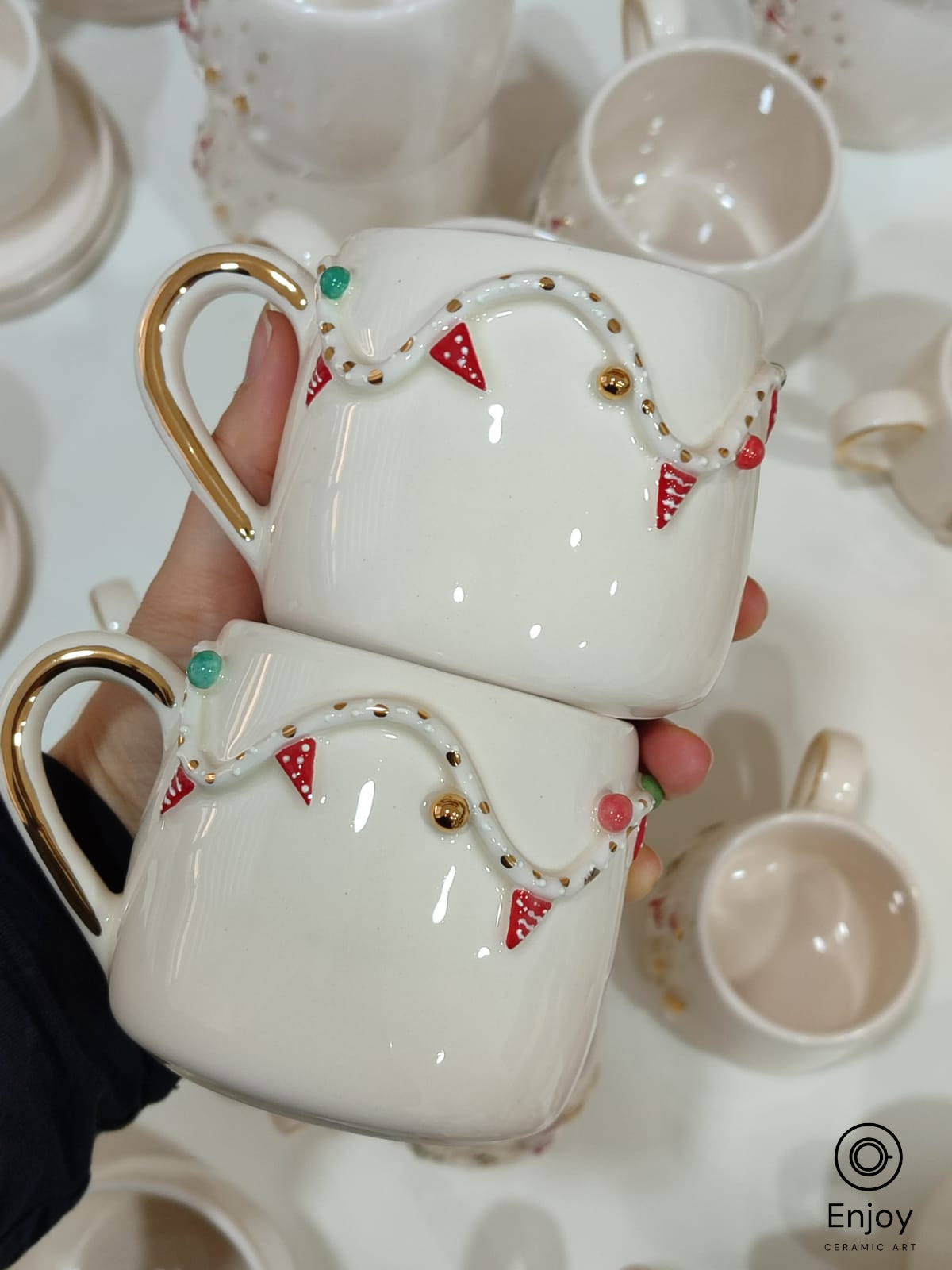 Two handmade ceramic mugs stacked together, featuring festive garland designs with red flags, gold accents, and colorful dots.