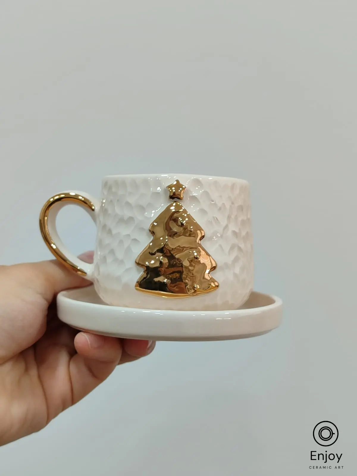 Hand holding a handmade ceramic espresso cup with a gold Christmas tree and star design, paired with a matching saucer.