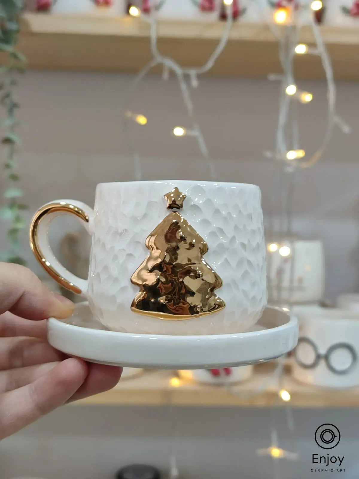 Hand holding a handmade ceramic espresso cup with a gold Christmas tree and star design, paired with a matching saucer.