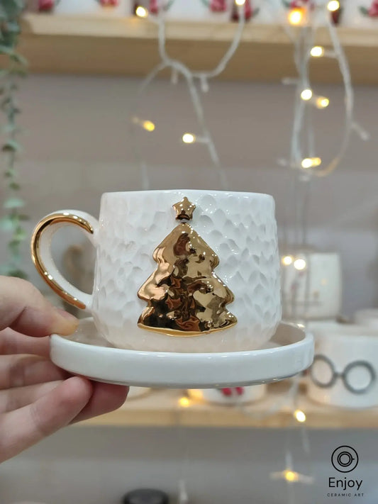 Hand holding a handmade ceramic espresso cup with a gold Christmas tree and star design, paired with a matching saucer.