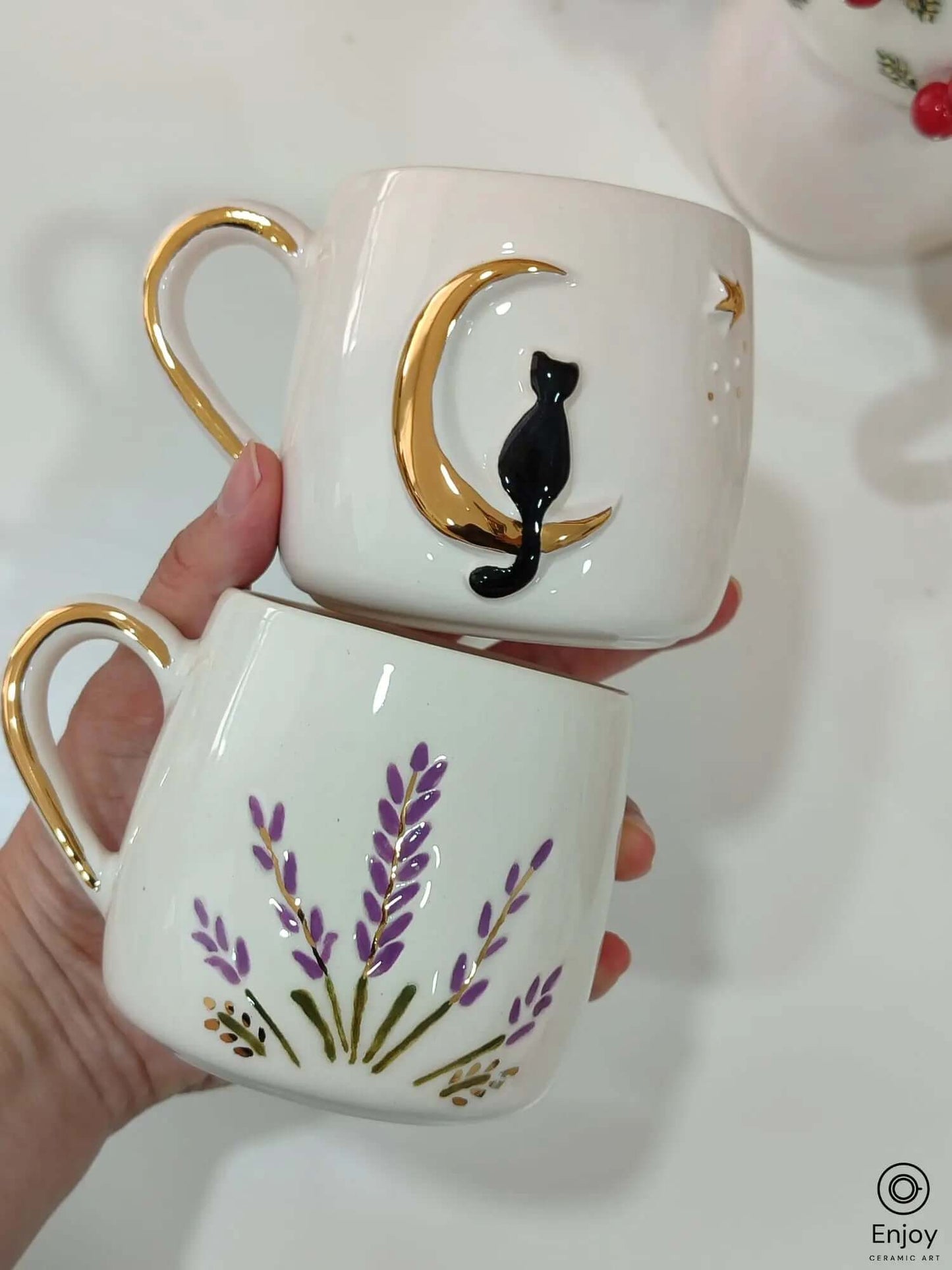 Handmade ceramic mug set featuring a black cat on a crescent moon and a lavender field design, both with gold handles and luxurious gold accents.