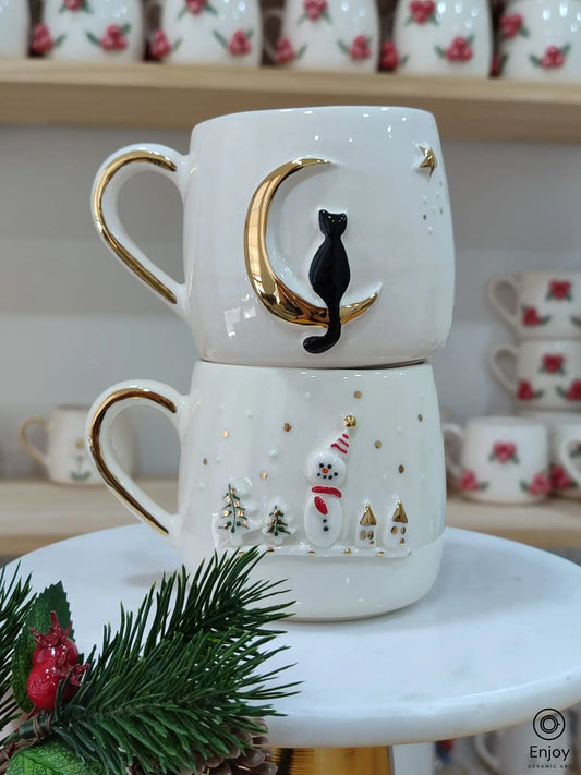 Handmade ceramic mug set featuring a black cat on a gold crescent moon and a snowman with red scarf and hat, accented with gold handles and Christmas designs.