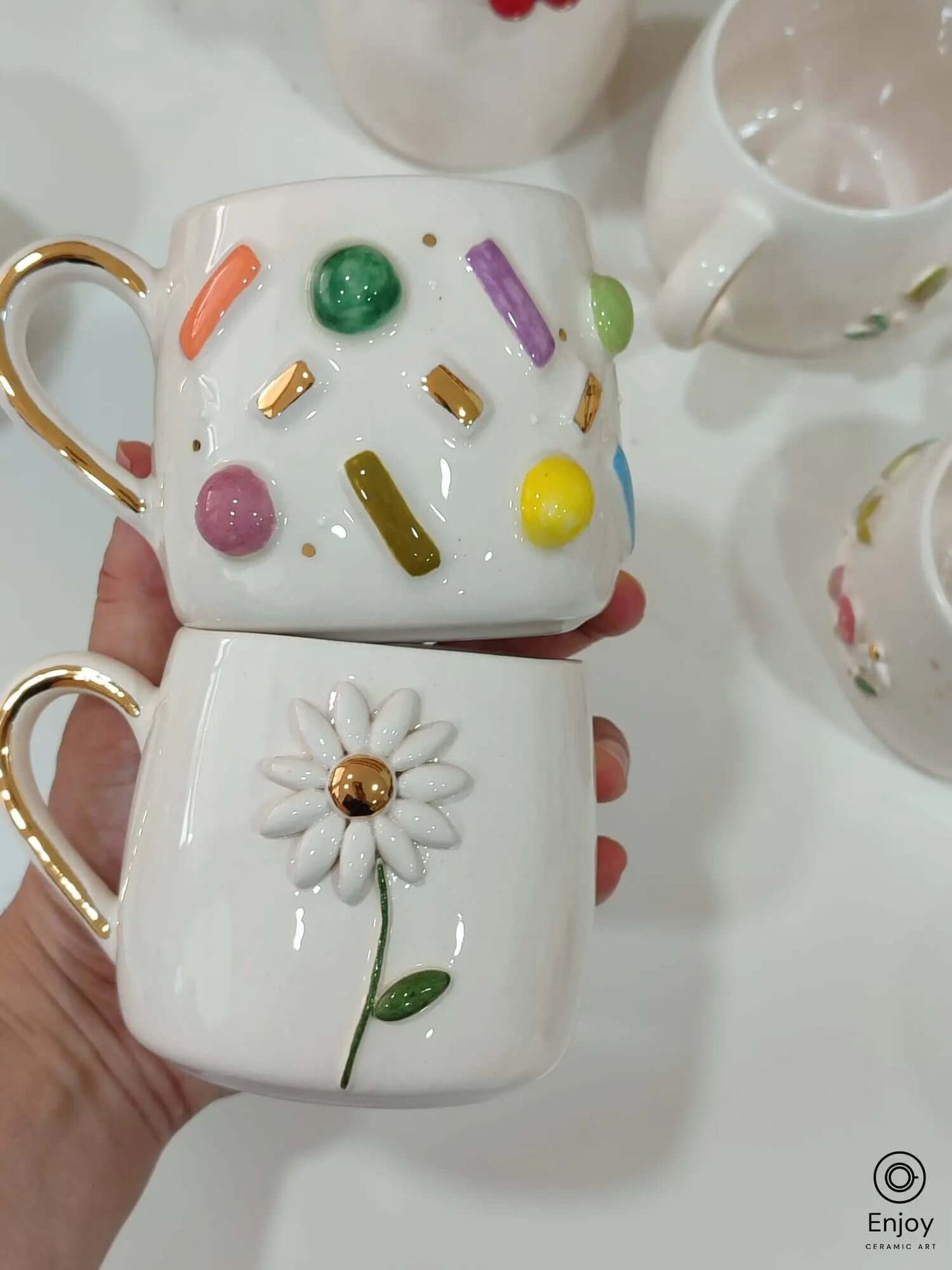 Handmade ceramic mug set with a 3D daisy flower and colorful sprinkle confetti design, both with gold handles and accents.