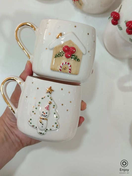 Handmade ceramic Christmas mug set featuring a 3D gingerbread house with candy cane details and a snowman inside a Christmas tree frame, both with gold accents and handles.