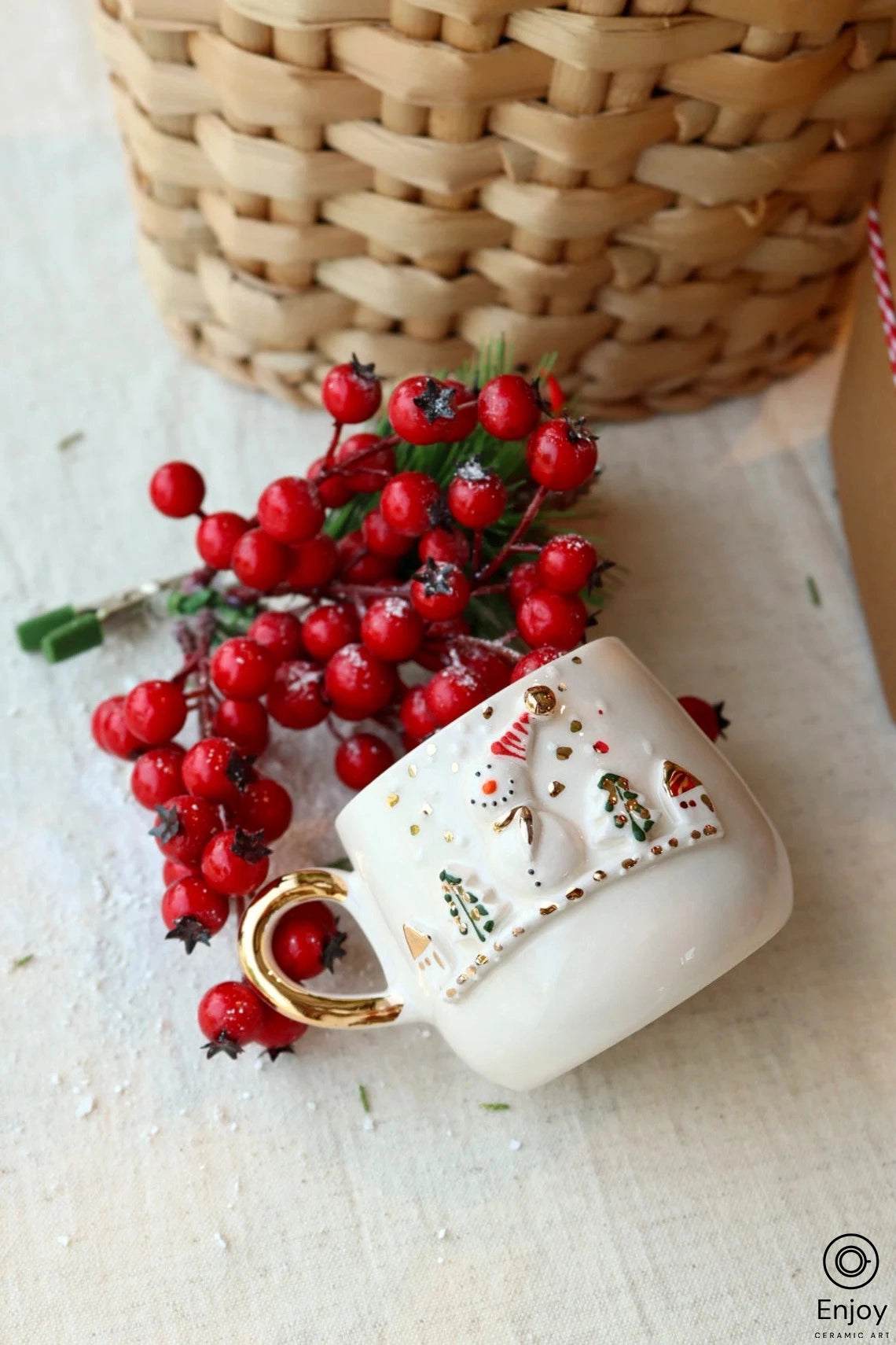 A festive espresso cup with golden rim and handle, adorned with a textured snowman and Christmas tree design, rests beside vibrant holly berries.