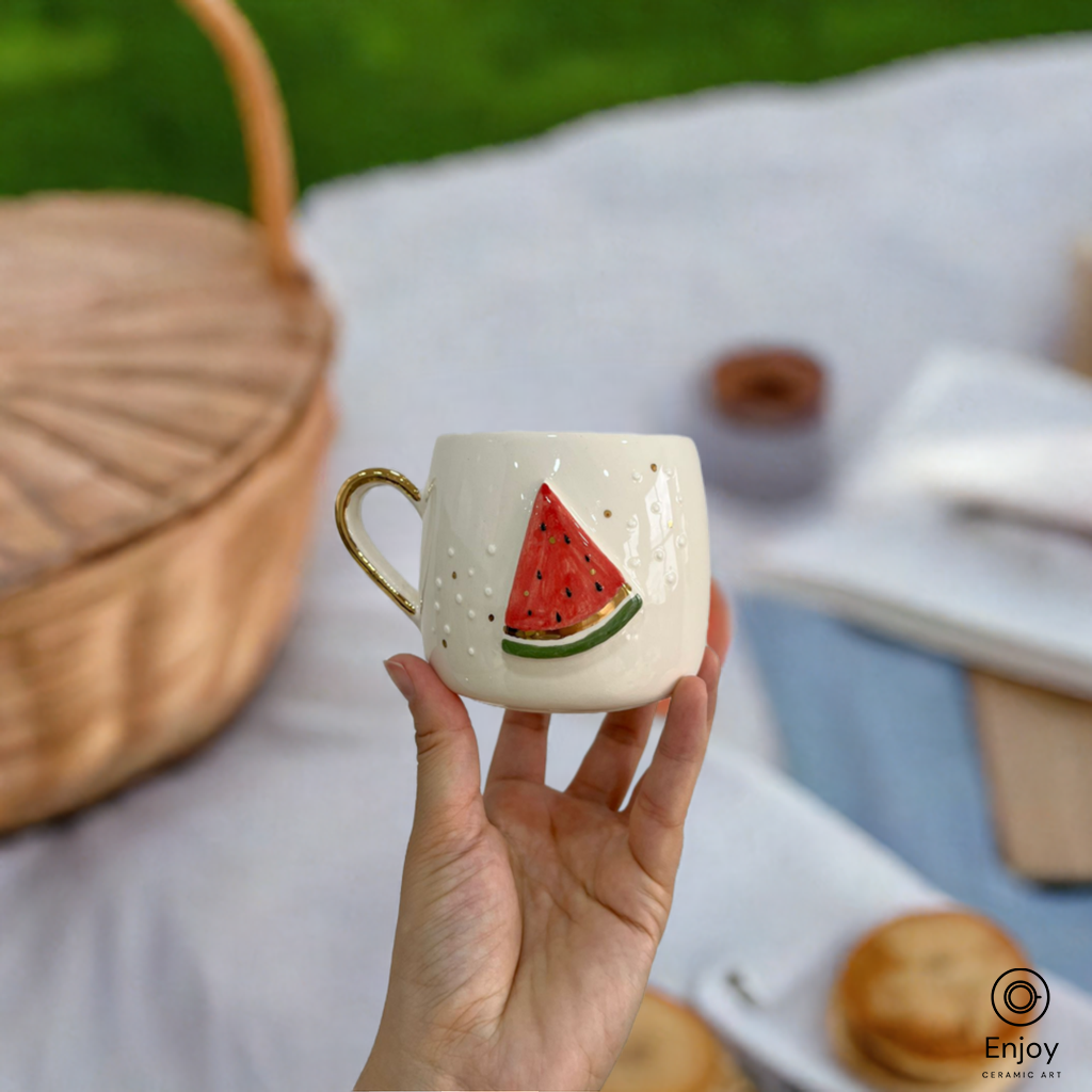 A handmade mug with a 3D watermelon design held in hand, against a cozy outdoor picnic backdrop.