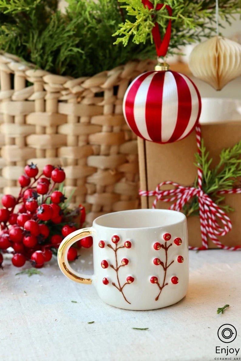 A festive white espresso cup with a gold handle and red berry branch designs, set against a backdrop of a woven basket, striped bauble, and greenery.