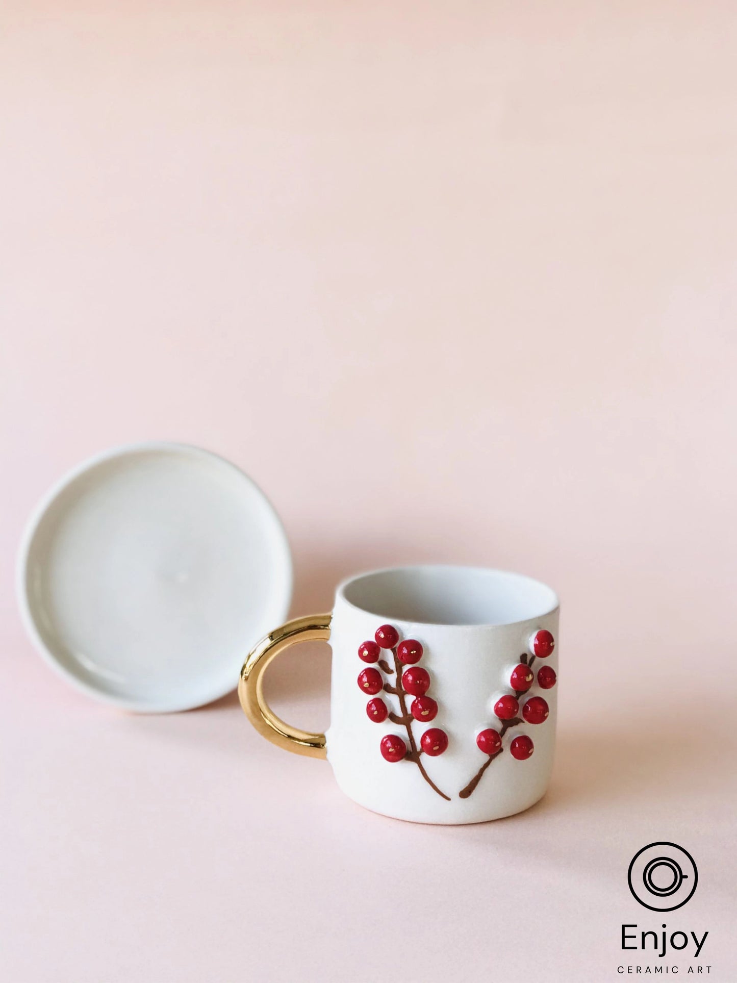 White ceramic espresso cup with a gold handle and raised red winterberry design, against a pale pink backdrop, by Enjoy Ceramic Art.