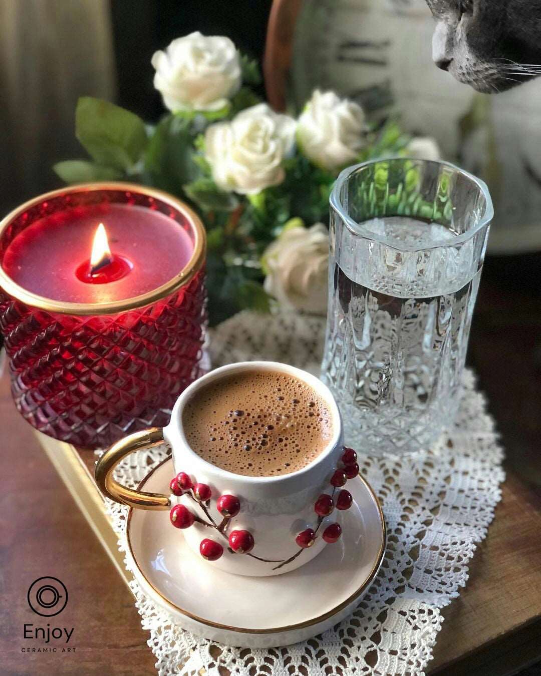 Aromatic espresso in a white and red winterberry-designed cup with a gold handle, on a saucer atop a lace doily, with a red candle, white roses, and a cat in the soft-focused background.