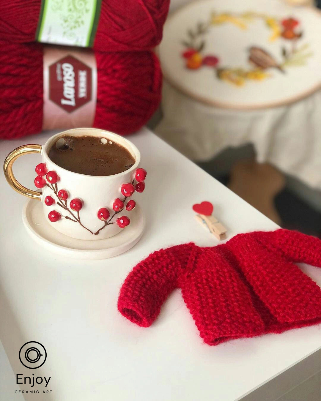 Handcrafted espresso cup with winterberry design and gold handle, filled with coffee, beside a red knitted cardigan and crafting supplies, evoking a warm, artistic atmosphere
