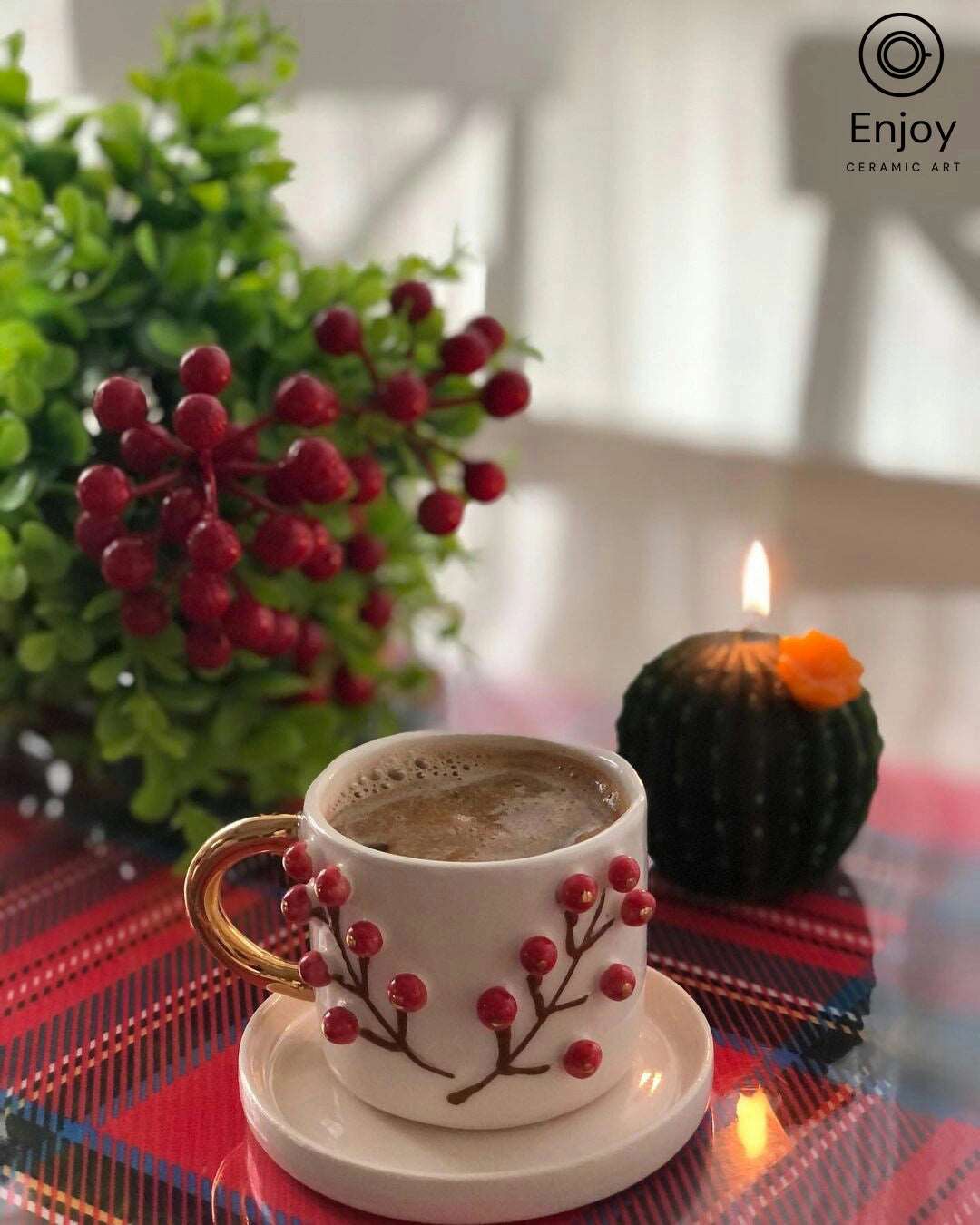 Frothy espresso in an artisanal white cup with a gold handle and winterberry motif, placed next to a green plant with red berries and a lit candle, creating a cozy ambiance.
