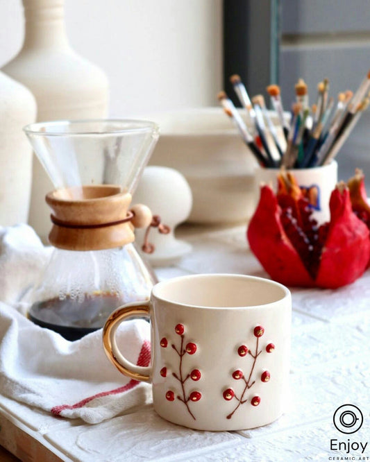 Artistic still-life of a white ceramic cup with red winterberry design and gold handle, next to a manual coffee dripper and paintbrushes, with a pomegranate section in the background.