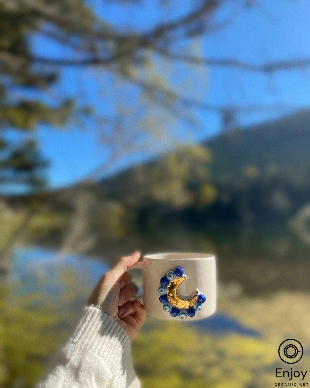 A hand holding a 'Blue Moon' ceramic mug with gold crescent and blue bead details, with a blurred natural landscape in the background, evoking a sense of warmth and tranquility.