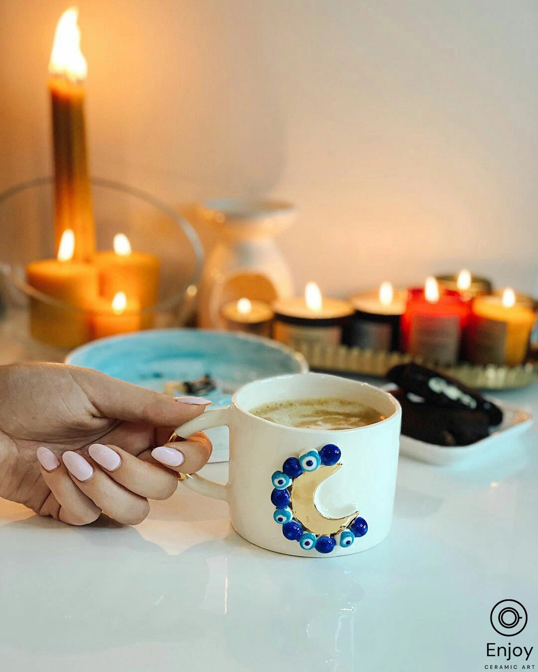 A hand holding a 'Blue Moon' ceramic mug with a golden crescent moon and blue evil eye beads, against a serene backdrop of soft candlelight.